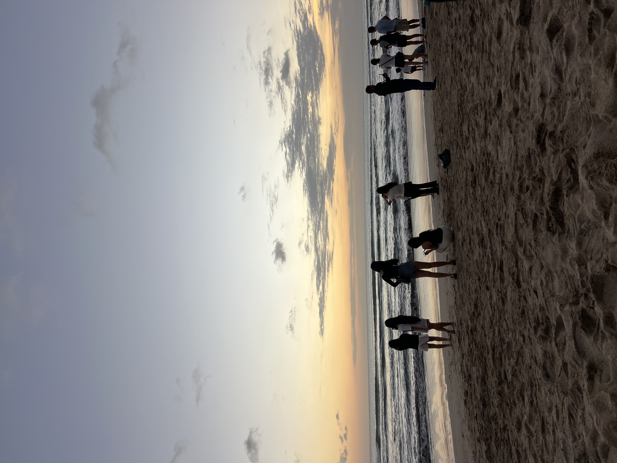 At sunset on Kuta Beach, Indonesia, a group of people stand on the sand and at the water's edge, silhouetted against a sky painted with hues of orange, yellow, and soft gray. Gentle waves roll onto the shore, reflecting the fading light. Several individuals are captured in candid moments: a woman stands with her back to the camera, hands on her hips; a man holds a phone, possibly taking a photo; a group of young women stand together, facing the sea; and a couple with a child appear to be enjoying the view. The sandy beach is textured with footprints and ripples, and a dark bag lies discarded near the water. The overall atmosphere is tranquil, capturing the serene beauty of a tropical evening.