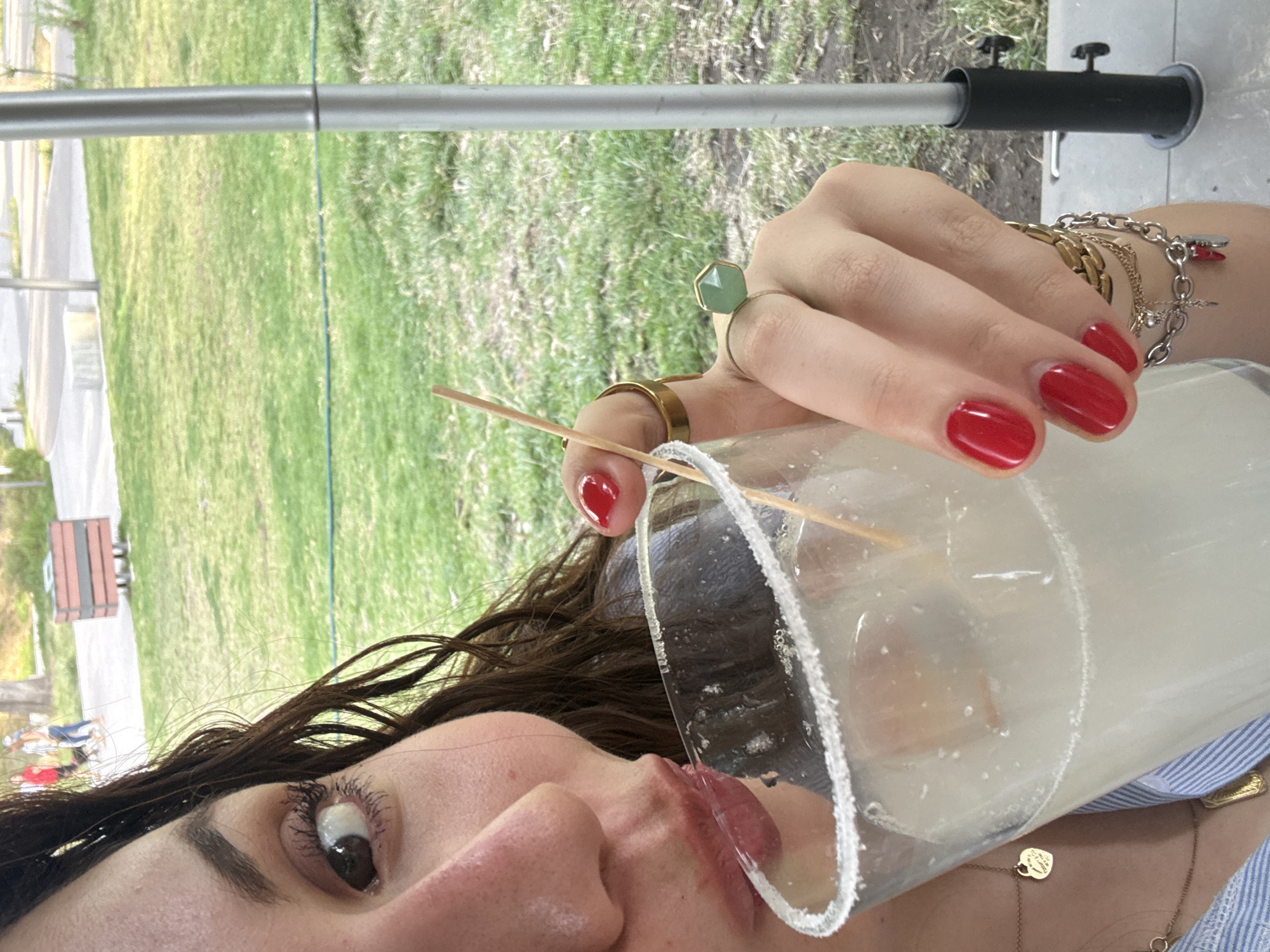 The image shows a woman with brown hair and red nail polish taking a drink from a glass rimmed with salt. She is wearing multiple rings and bracelets. A wooden stick protrudes from the drink. The background includes green grass and a metal structure. Given the presence of lush greenery and metal structures, it's plausible, but not definitively confirmed without additional context, that the location might be a park or outdoor area in Mexico City, Mexico.