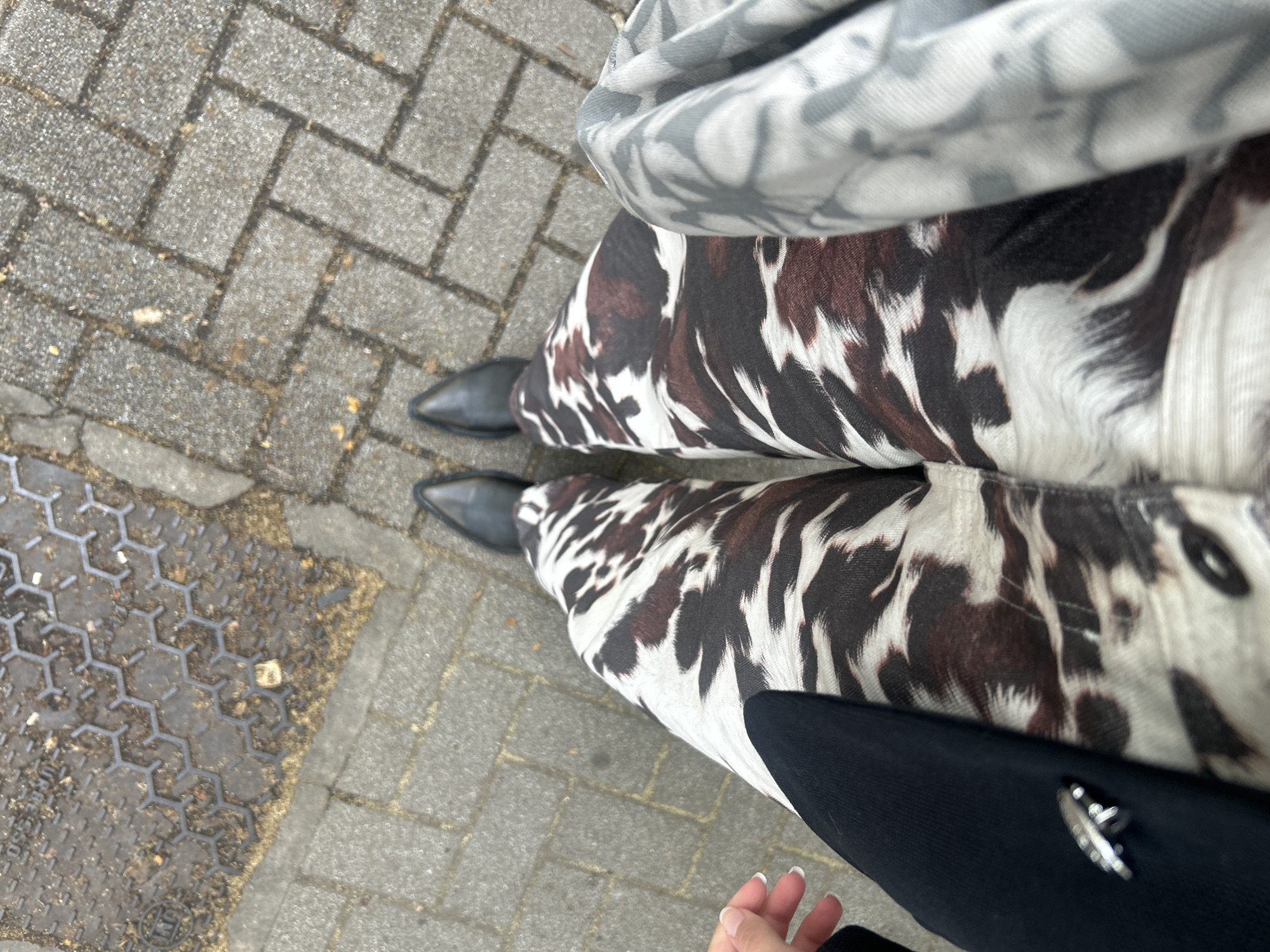 The image shows a person standing on a paved sidewalk, wearing cow-print pants and black pointed-toe boots. A grey patterned scarf is draped on the upper body, and part of a black jacket or coat is visible on the right side of the image, with a silver leaf pin on it. The sidewalk is made of gray rectangular pavers, with a section of a hexagonal patterned drain cover visible on the left side. The location is Milan, Italy.