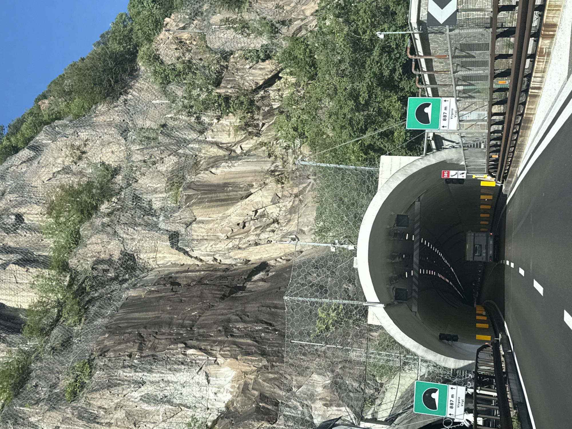 The image shows a view from a vehicle on a road approaching a tunnel entrance. The tunnel is carved into a steep, rocky mountainside with a protective mesh covering the rock face. Green traffic signs with a tunnel symbol and distance markings are visible to the left and right of the road. The road is paved and has white lane markings. Yellow delineators line the sides of the road. Inside the tunnel, a recreational vehicle can be seen. The sky is clear and blue.
