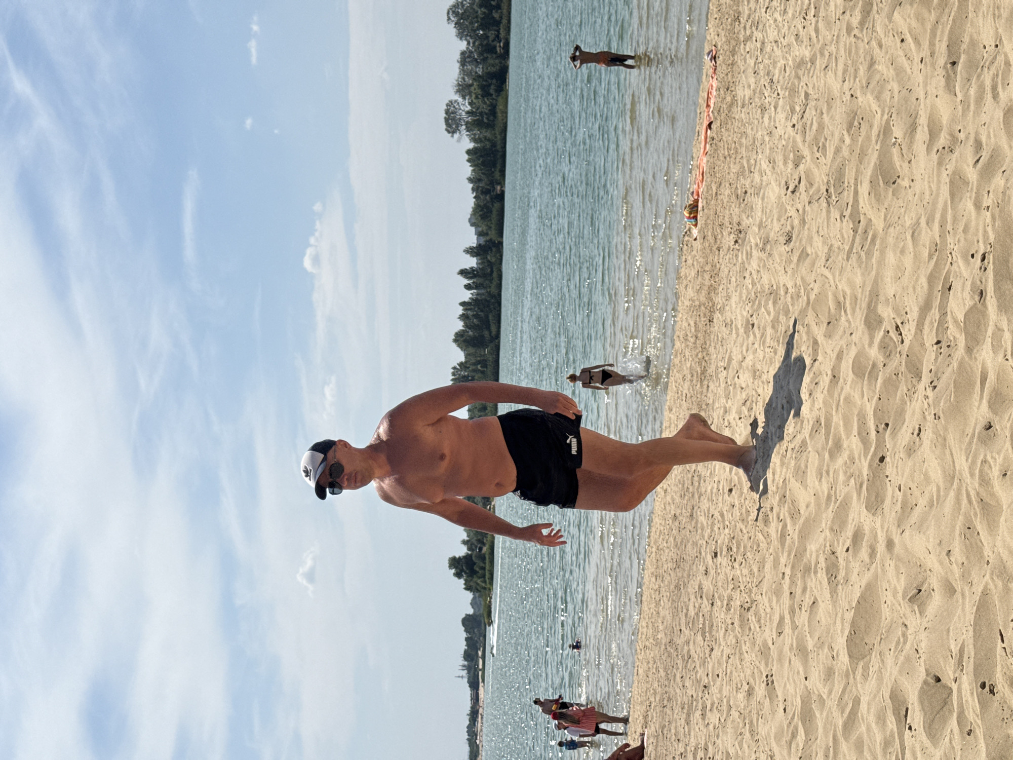 A man wearing black Puma swim trunks, a white and black baseball cap, and sunglasses walks on a sandy beach. He is in the foreground of the image, facing left. The beach is adjacent to a body of water that appears to be a lake or reservoir, with a treeline in the distance. Several other people are visible in the water and on the beach, some swimming and others relaxing. The sky is blue with scattered white clouds. The location is Yelyzavetivka, Ukraine.