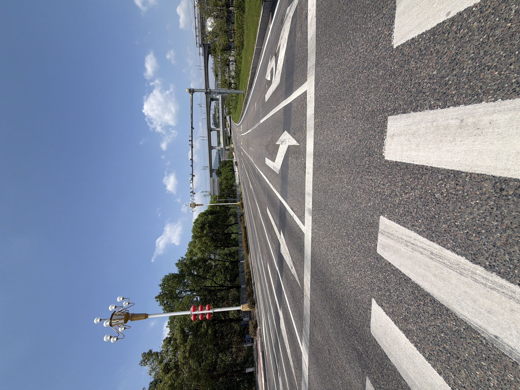This is a low-angle, eye-level, wide shot of a road in Jiangjin District, Chongqing, China. The road is paved with asphalt and marked with white directional arrows and crosswalk lines. The sky is clear blue with scattered white clouds. Trees line the left side of the road, and a pedestrian overpass or bridge is visible in the distance on the right, with train tracks running along it. A traditional-style street lamp with multiple globes stands on the left side of the road, and a traffic light with red lights is attached to it. The scene appears to be a public road, possibly leading into or out of a more urban area, given the infrastructure in the background. There are no people or vehicles visible in the frame, giving the impression of a quiet, possibly early morning or late afternoon scene.