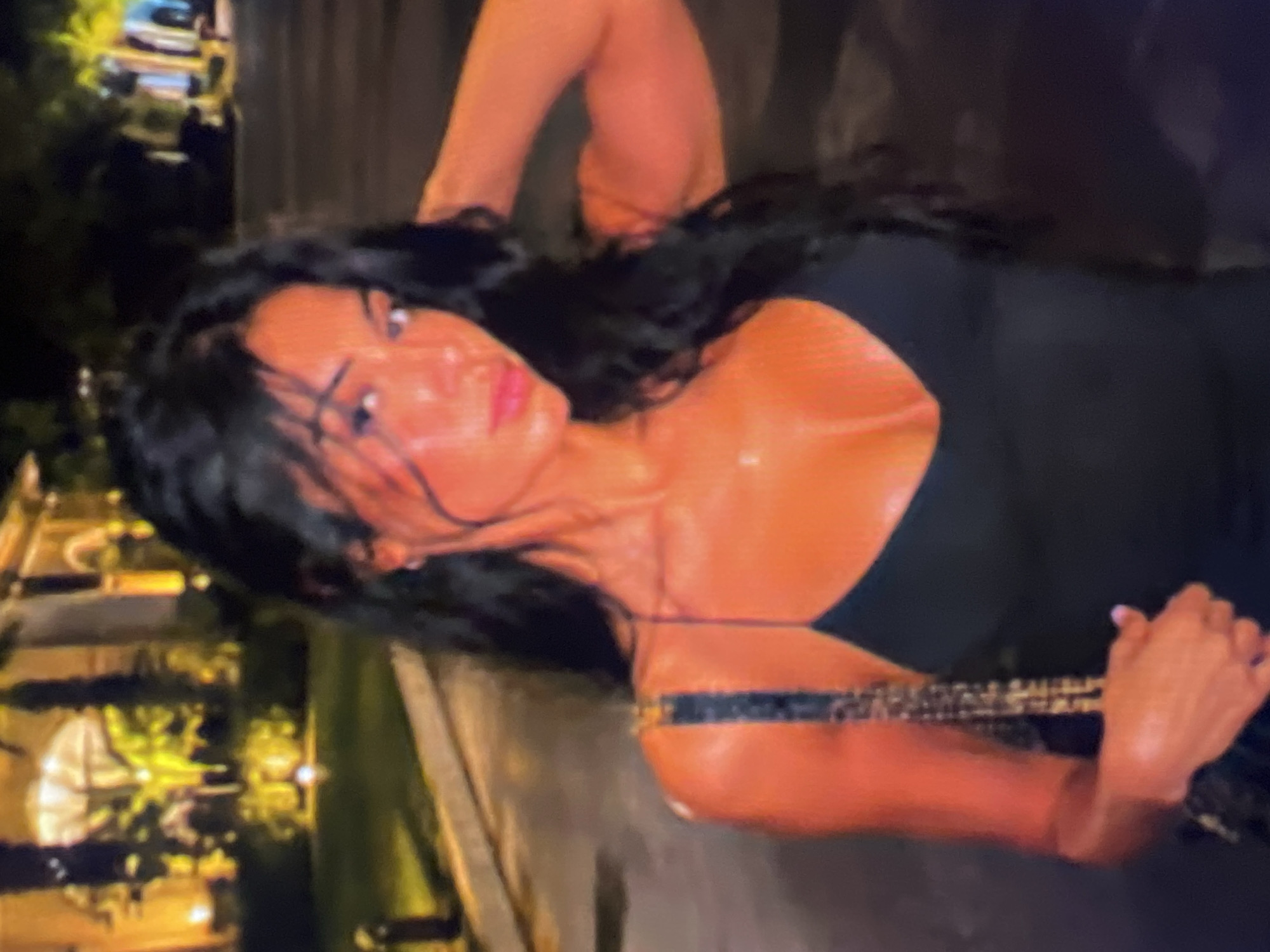 An outdoor, nighttime, medium shot captures a young woman with long, dark, wavy hair. She is wearing a black spaghetti strap dress and a beaded purse. The woman is positioned in the foreground, facing the camera with her head tilted slightly. Her skin appears bronzed and her lips are painted red. She has her right arm raised and is touching her hair. In the background, a building with illuminated arched windows and palm trees are visible. The lighting is warm and orange, suggesting artificial lighting from the building and possibly streetlights. The ground appears wet, indicating it may have rained recently.