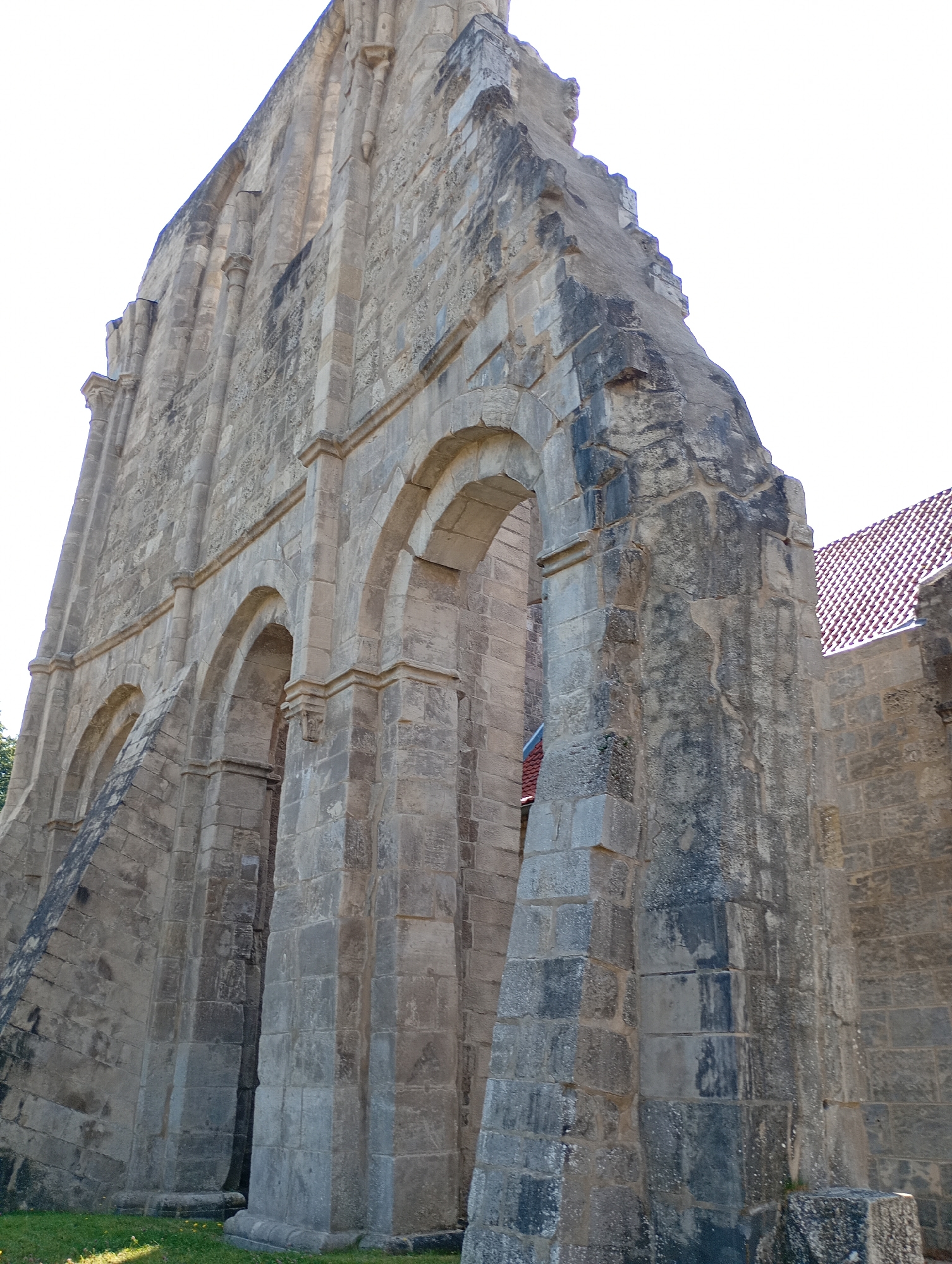 The image shows a low-angle exterior view of the ruins of a large stone structure, likely a church or abbey, against a bright, overcast sky. The dominant feature is a massive stone wall constructed from large, rectangular blocks of weathered grey stone. The wall is angled, rising sharply to the left and extending towards the right.
Several prominent arched openings pierce the wall, supported by thick, pilaster-like columns. The arches appear to be Romanesque in style. The stonework shows signs of age, with patches of dark staining and erosion. The top edge of the wall is jagged and broken, indicating it is a ruin.
To the right of the main structure, a section of a building with a red-tiled roof is visible. This suggests that part of the original complex may still be intact or has been partially restored. The ground in the foreground is covered in green grass with scattered yellow flowers, indicating an outdoor setting.
This image depicts the ruins of the Walkenried Abbey in Walkenried, Germany. The abbey is a significant historical site, known for its impressive Gothic and Romanesque architecture. The ruins are a testament to its long history, which includes periods of construction, damage, and preservation.
