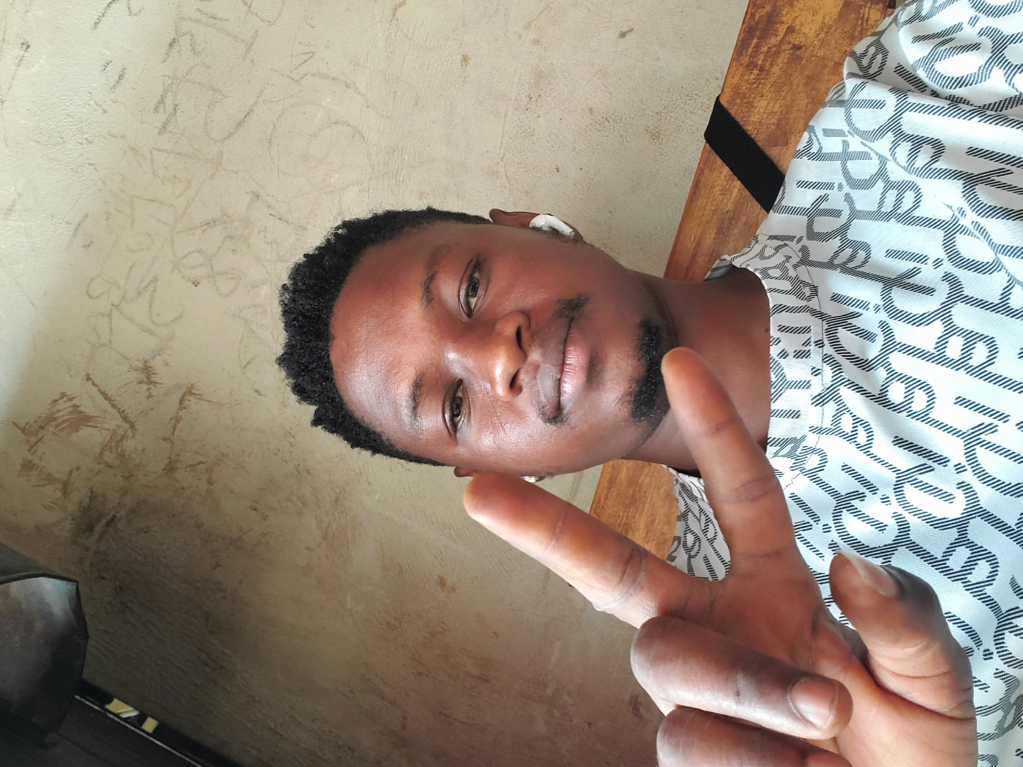 A close-up, slightly tilted portrait shows a young Black man, likely in Mararaba, Nigeria, in an indoor setting. He has short, dark curly hair and a small, neat goatee. A white earbud is visible in his left ear. He is wearing a white short-sleeved shirt adorned with a repeating pattern of black geometric designs and parallel lines.
The man looks directly at the camera with a calm, neutral expression. His right hand is raised in the foreground, making a peace sign (V-sign) with his index and middle fingers extended.
The background features a light beige or cream-colored wall that appears somewhat worn, with faint, scratched-in markings and scribbles. These markings are not clearly discernible as legible text or symbols. A dark wooden surface, possibly a piece of furniture, is visible to the right of the man. The lighting is soft and even, suggesting an unstated time of day.