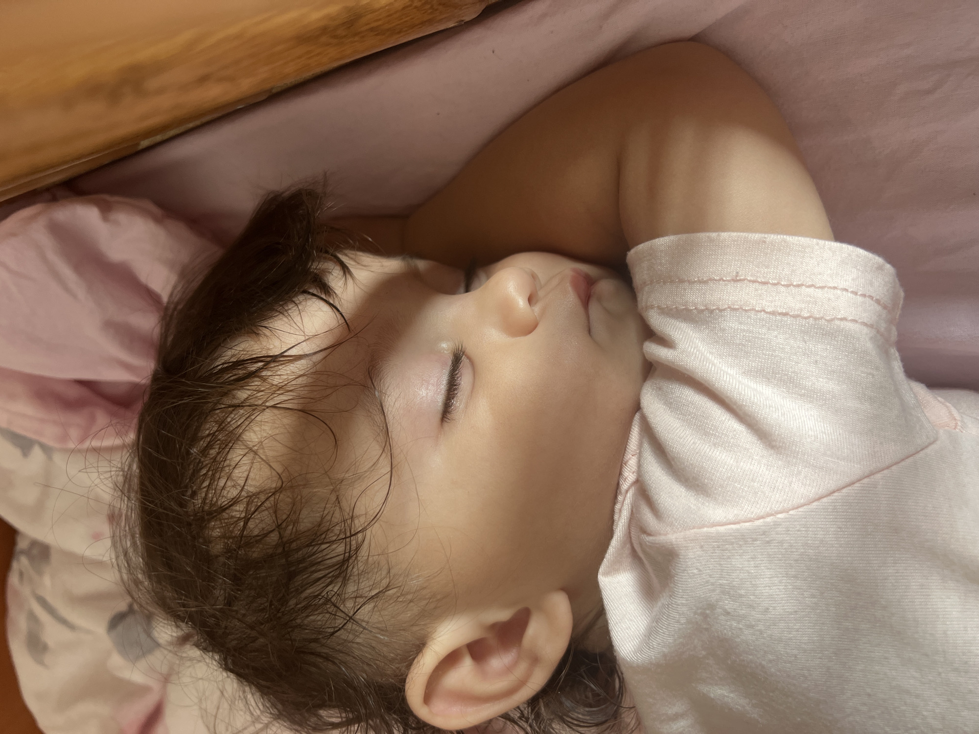 A close-up, eye-level shot shows a young child sleeping on their side, facing right, in what appears to be a bed. The child is wearing a light pink t-shirt. Their head rests on their arm, which is bent at the elbow, creating a pillow. Dark, wispy hair is spread around the child's head. The background consists of a pink surface, likely a sheet or blanket, with some of it bunched up behind the child's head. The lighting suggests an indoor setting. There is no information available to determine the location in the city of Elena, Bulgaria.