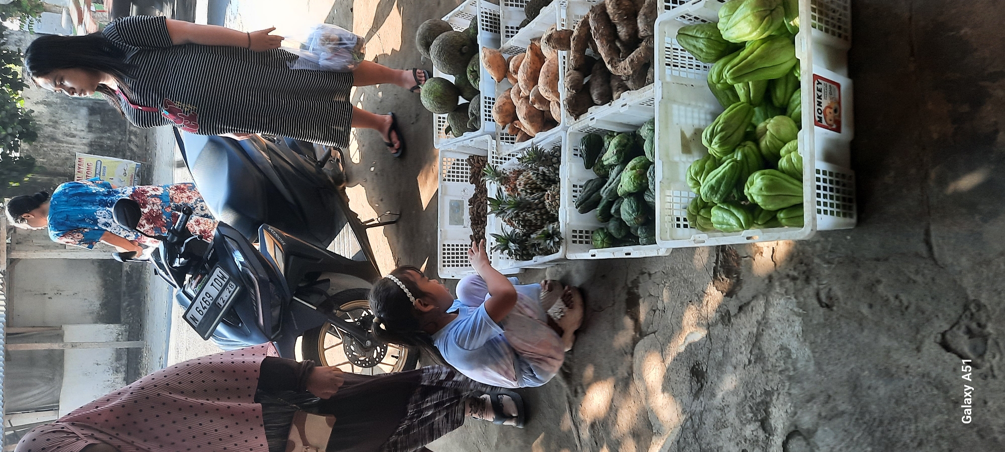 In Selopuro, Indonesia, a young girl with dark hair, wearing a light blue t-shirt and light-colored pants, kneels on a concrete surface, reaching into a white plastic crate filled with small, brown, elongated fruits, possibly tamarind. She is looking down at the fruits and has a silver headband on. Beside her, a motorcycle is parked. Behind the girl, a woman in a striped black and white dress stands, holding a clear plastic bag with produce. Further behind her, another woman in a patterned blue and white dress stands near the motorcycle, with her back to the camera. In the background, a weathered building with a corrugated roof and a sign with Indonesian text is visible. In front of the crates, there are other white plastic crates filled with various produce, including pineapples, green squash, and sweet potatoes. The ground is a textured concrete with patches of sunlight and shadow. The overall scene depicts a street-side market setting.