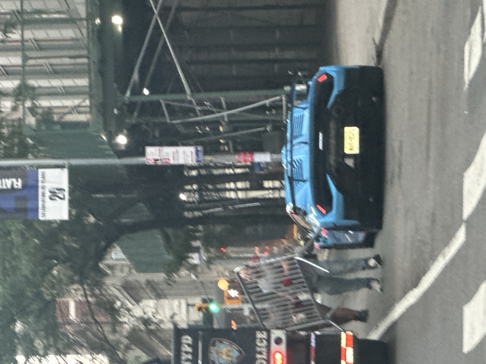 In New York City, a bright blue Lamborghini is parked on a street next to an NYPD vehicle. A person in gray pants and a white shirt is pushing a metal barricade towards the Lamborghini. Another person, wearing brown pants and black shoes, stands behind the barricade. The scene appears to be a street in the city, with buildings and trees visible in the background, suggesting an urban environment.