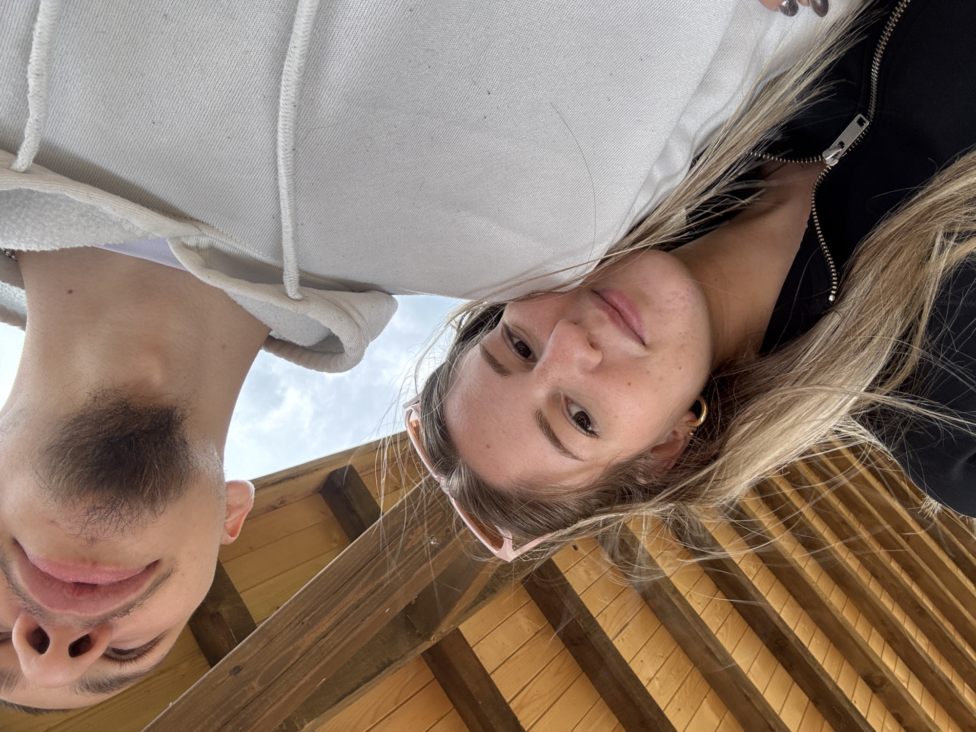 The image shows a selfie taken from a low angle, presumably by one of the two people in the frame. The location appears to be outdoors, possibly on a wooden deck or patio with a slatted wood ceiling. The photo includes two people: a man on the left and a woman on the right. The man has a beard, and the woman is wearing sunglasses perched on her head and gold hoop earrings. The woman appears to be wearing a black zip-up jacket. The sky is partially visible in the background, indicating it is daytime. The photo was taken at Цигов чарк, Bulgaria.