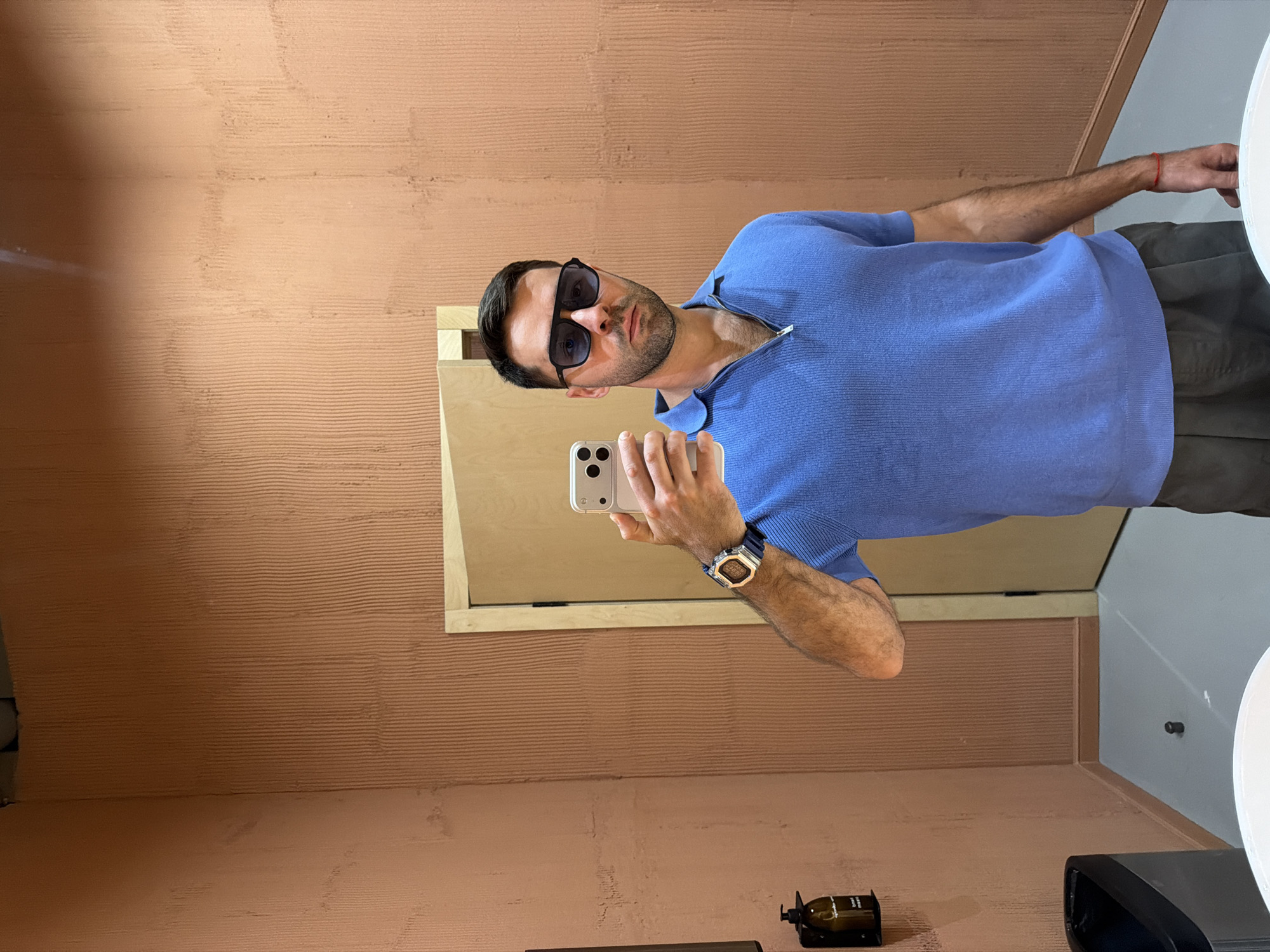 A man with dark hair and a beard, wearing sunglasses and a blue shirt, is taking a selfie in a restroom. He is holding a white smartphone. He is wearing a watch on his left wrist. The restroom has textured, peach-colored walls. A mirror, a soap dispenser, and a hand dryer are visible. The exact location in Krasnoyarsk, Russia, cannot be determined from the image.