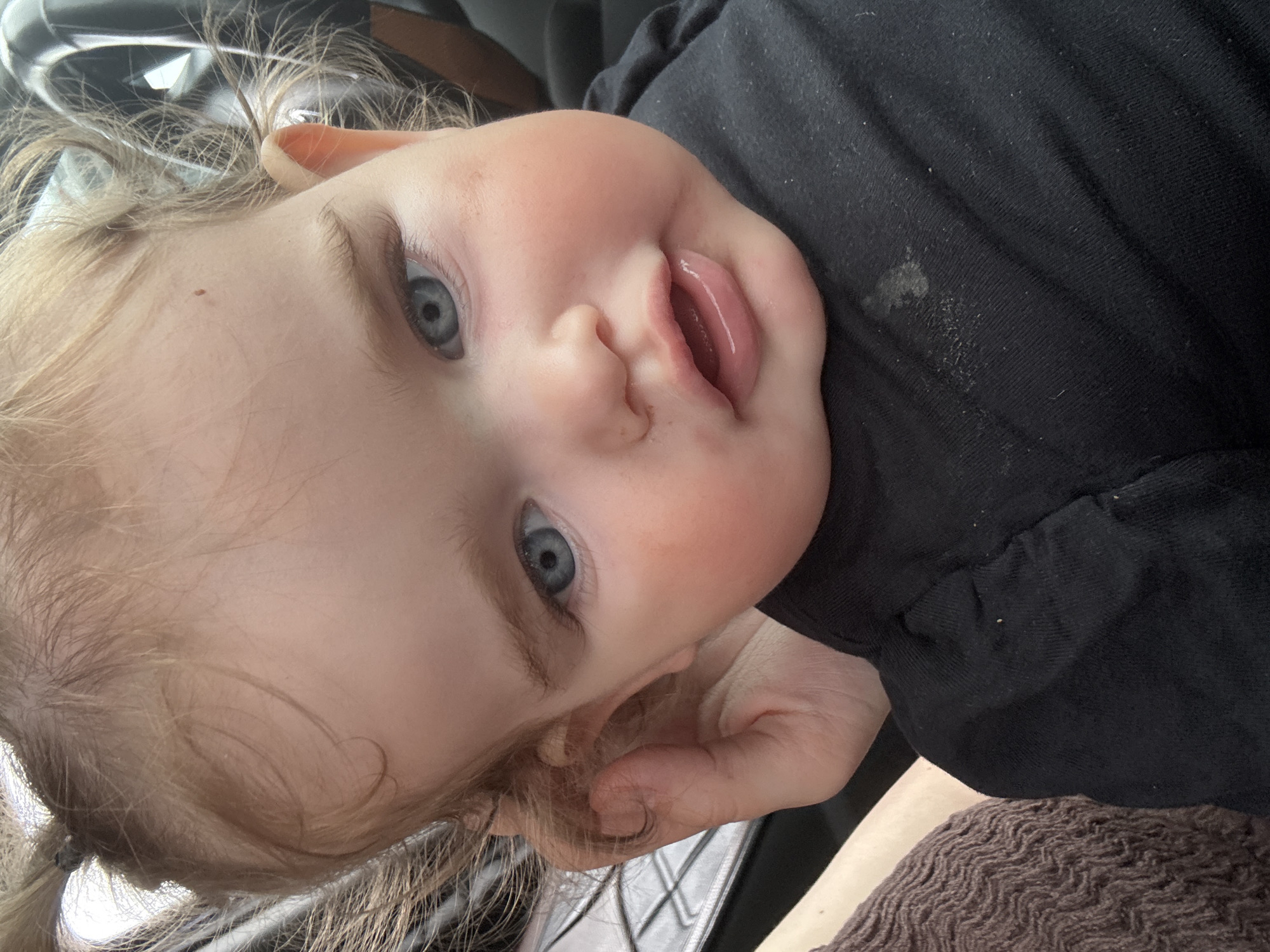 This is a close-up, high-angle, vertical, eye-level shot of a young child's face, likely a toddler, looking upwards. The child has fair hair, possibly blonde, which is a bit messy and has a slight curl. One of the child's eyes is visible, showing a clear blue iris. The child's lips are slightly parted, with the tongue visible. The child has rosy cheeks, typical of a young child. The child is wearing a black garment, which appears to be a shirt or dress, with some light-colored smudges on it. Part of an adult's hand is visible near the child's chin, gently touching the child's face. The background is blurred but suggests an indoor setting, possibly a car, with hints of a car seat and window. The lighting is bright, indicating daylight. The child's expression appears curious and engaged. No text is visible in the image. The location context is Koblenz, Germany.