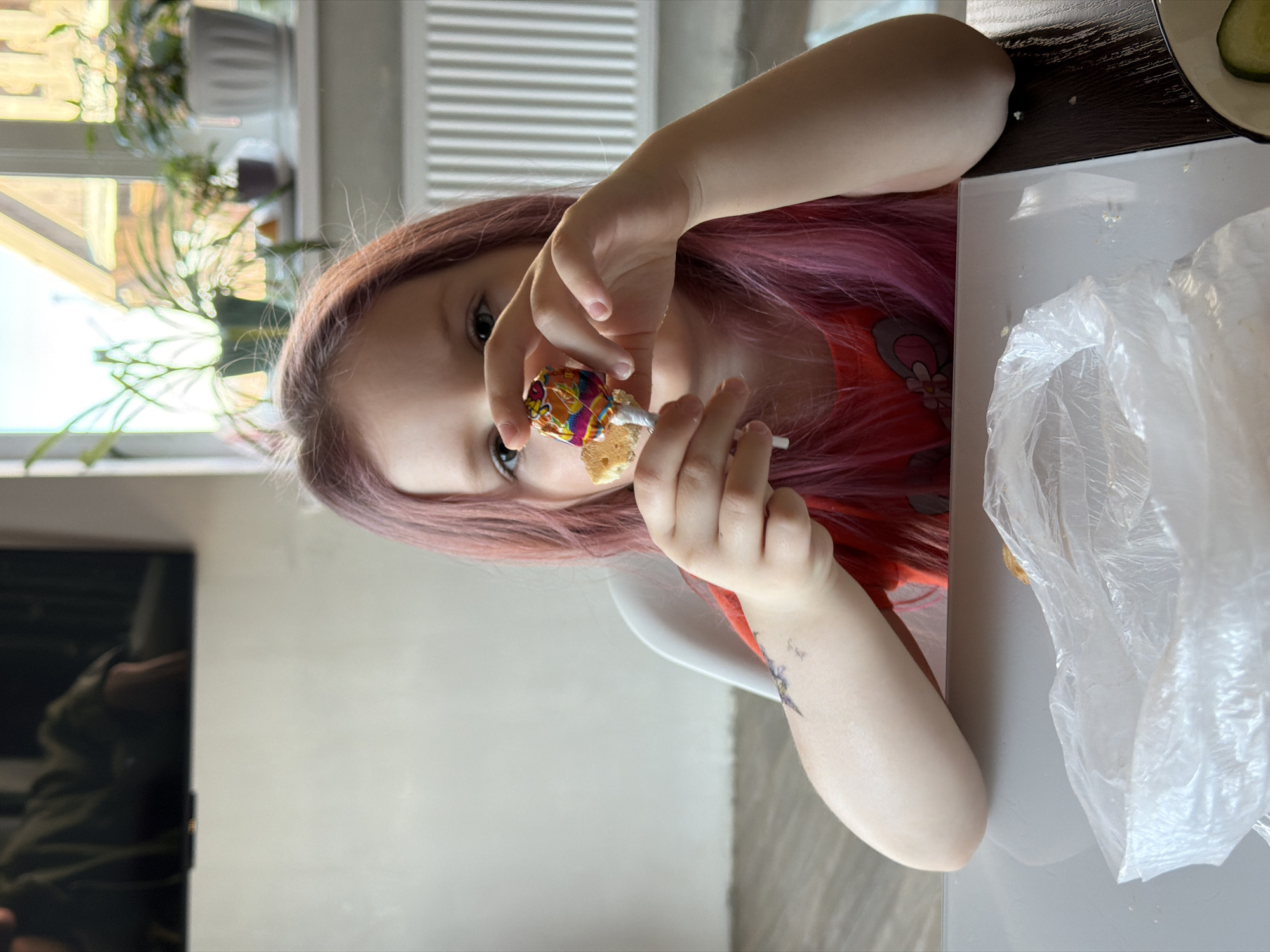 A young girl with long, pink-tinted hair is seated at a table, holding a lollipop in her hands. The girl is wearing an orange shirt and has a tattoo on her left arm. The table is white and cluttered with a plastic bag. Behind the girl, there is a window with plants and a radiator. To the left, there is a blurry image of a television. The location is in the city of Chaikovsky, Russia.