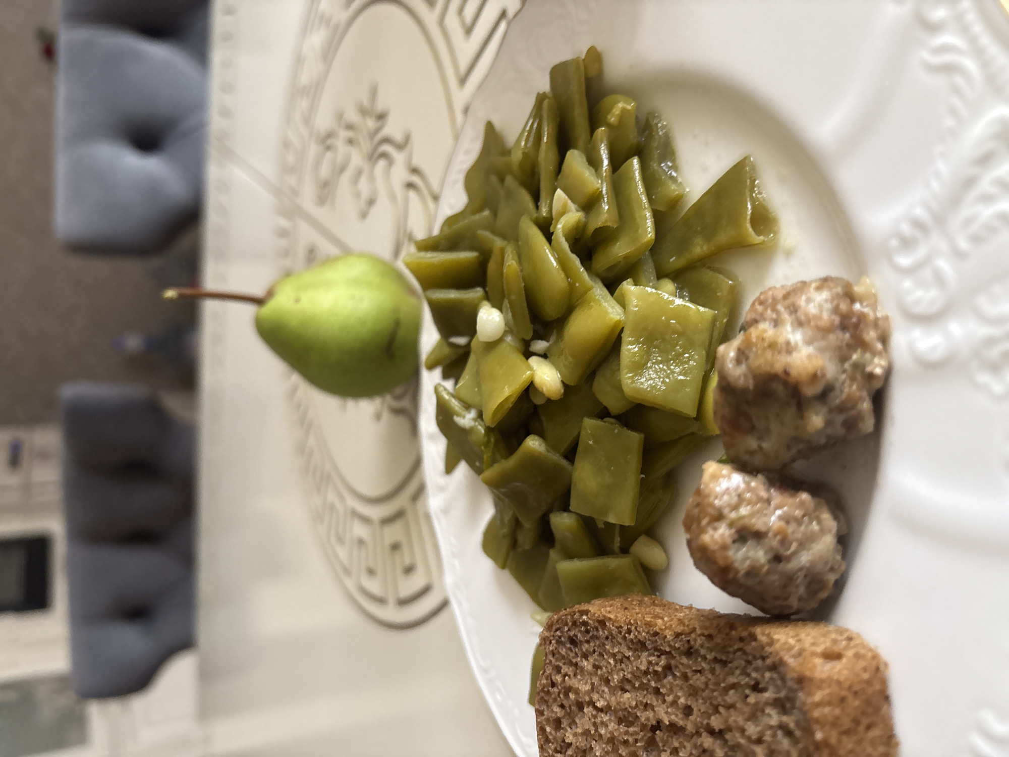 On a white, ornate plate, a meal is presented, consisting of cooked green beans, two small meatballs, and a slice of dark bread. In the background, a green pear stands on a table with a patterned surface, beyond which two plush, dark gray chairs are visible, suggesting an indoor dining setting. The scene appears to be a still life of a meal, with no people or actions depicted. There is no information in the image to suggest a location in the city of Georgiyevsk, Russia.