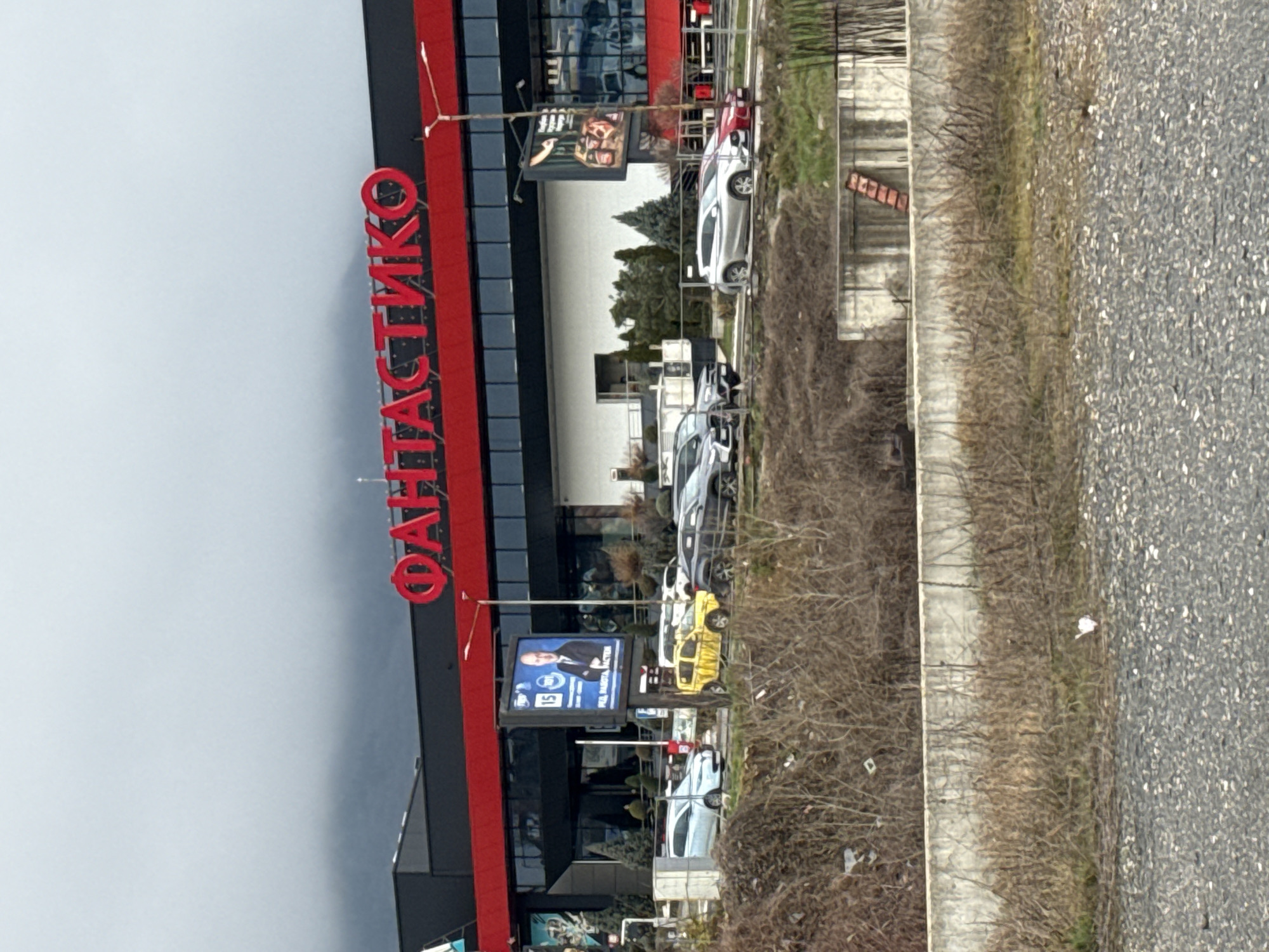 The image shows a view of a Fantastico supermarket in Sofia, Bulgaria. The large red Fantastico sign is prominently displayed on the building's facade. A row of parked cars is visible in front of the supermarket. A billboard displaying what appears to be a political advertisement is also visible. The image is taken from a slightly elevated position, looking towards the supermarket. The weather appears overcast. A sidewalk and roadway are to the right.