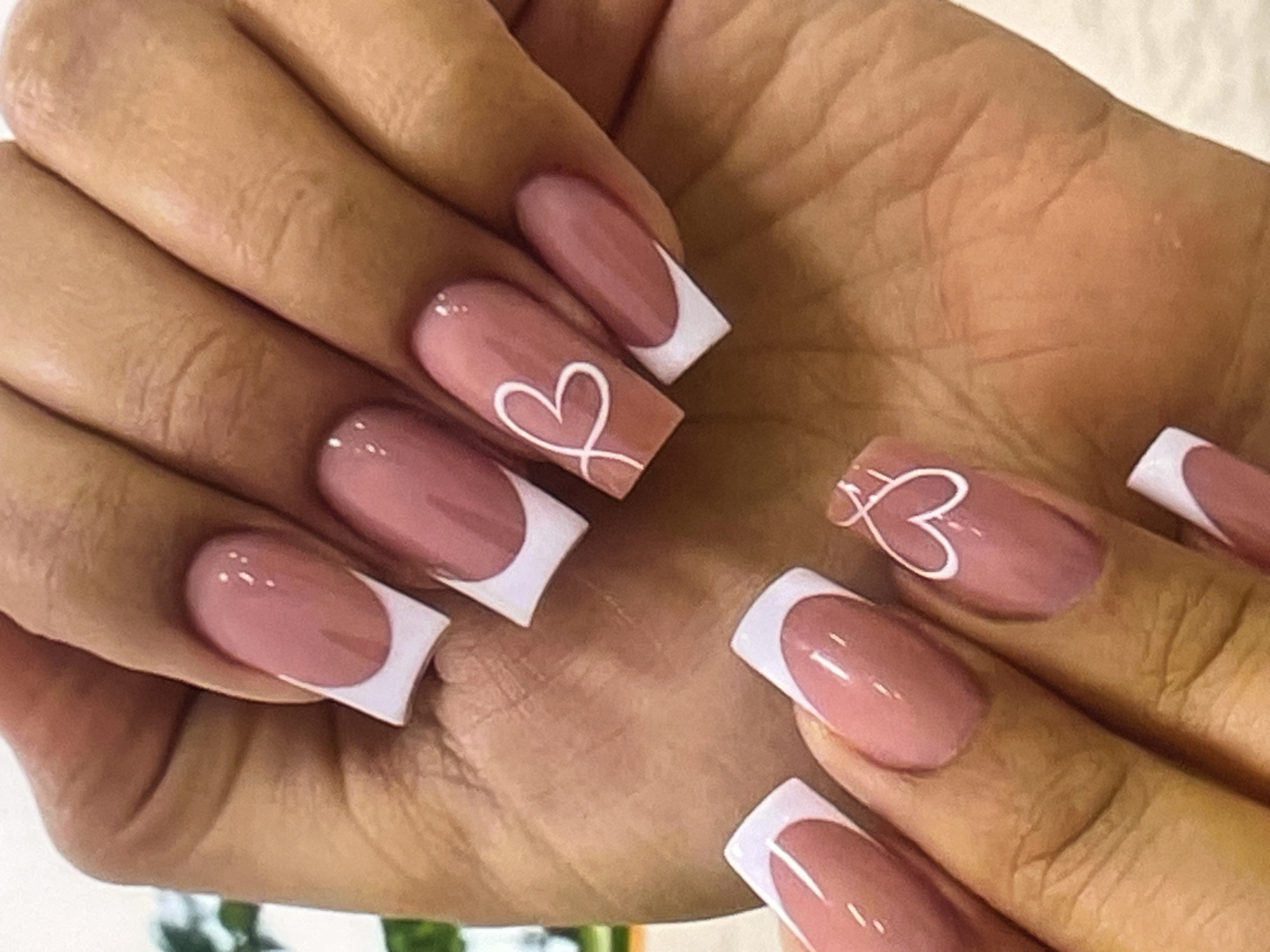 A close-up, overhead shot shows a person's hands displaying a French manicure with pink polish and white tips. The nails are square-shaped with rounded corners. Two nails on the right hand feature a white, abstract heart design. The hands are resting on a light-colored, textured surface, possibly a table or fabric. The background is softly blurred, with hints of green foliage suggesting an outdoor or plant-filled indoor setting.
