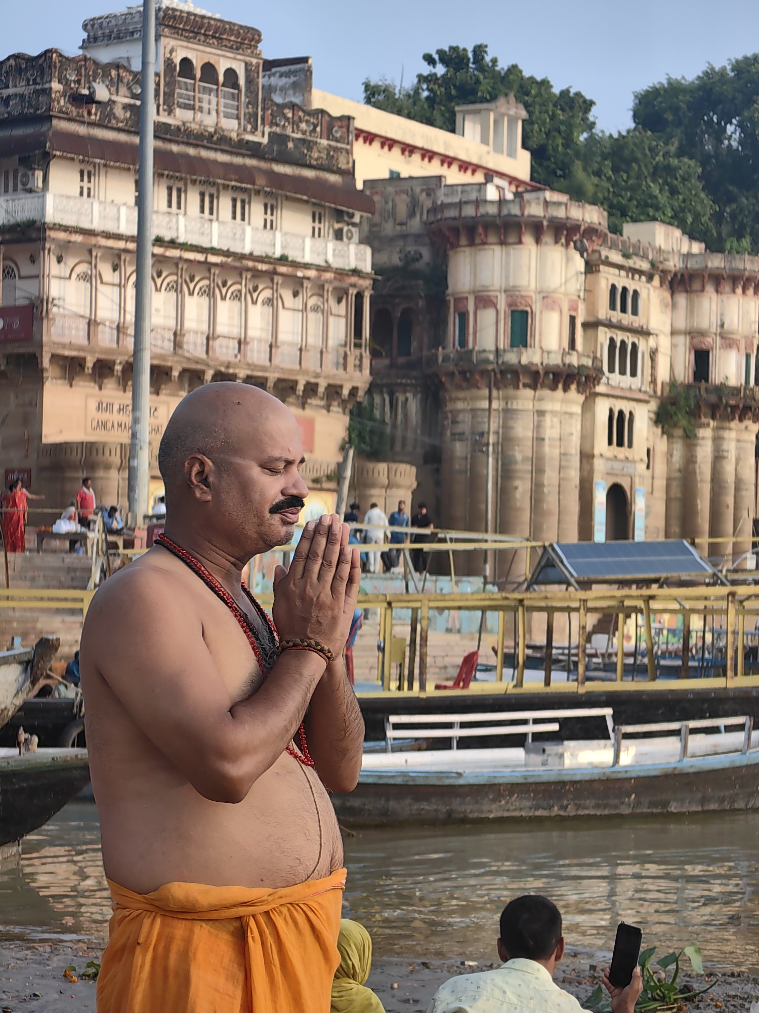 An outdoor, medium shot captures a bald, shirtless man with a mustache in profile, his hands clasped in prayer. He wears an orange dhoti and a beaded necklace, suggesting a religious or spiritual context, likely on the ghats of Varanasi, India, given the architectural style of the buildings in the background. The buildings, characterized by intricate carvings and multiple balconies, are a prominent feature of the Varanasi skyline. In the distance, a yellow railing separates the ghat from a body of water, on which a boat is docked. Other figures are visible in the background, some walking and others seated near the water's edge. The scene appears to be set in the early morning or late evening, indicated by the soft lighting and clear sky.