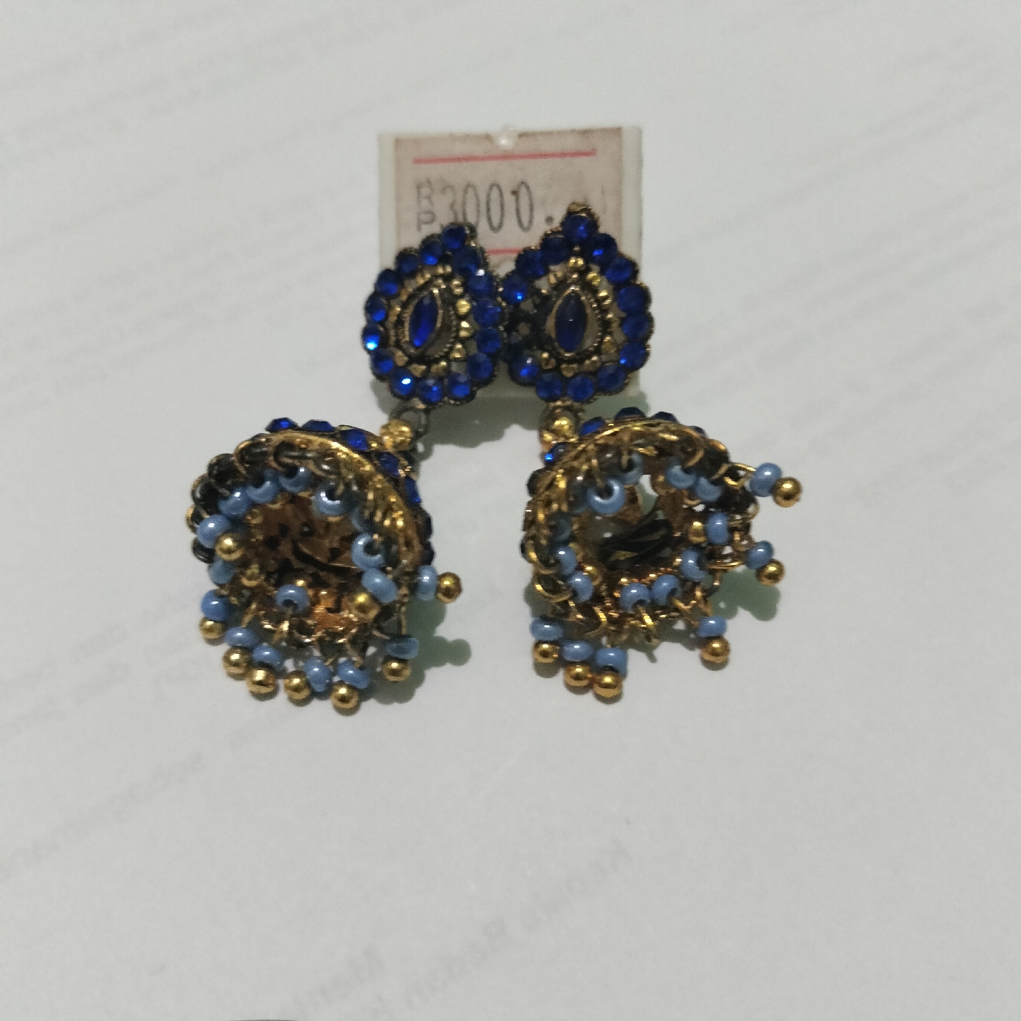 Two ornate jhumka earrings with blue stones and gold accents are displayed on a white surface. A price tag showing "3000" is visible behind the earrings. The earrings are of traditional Indian design. The background appears to be a plain white surface, possibly a table or paper. There are no people or other objects in the immediate vicinity. The image does not provide any information that could be used to determine the location in the city of Tangerang, Indonesia.