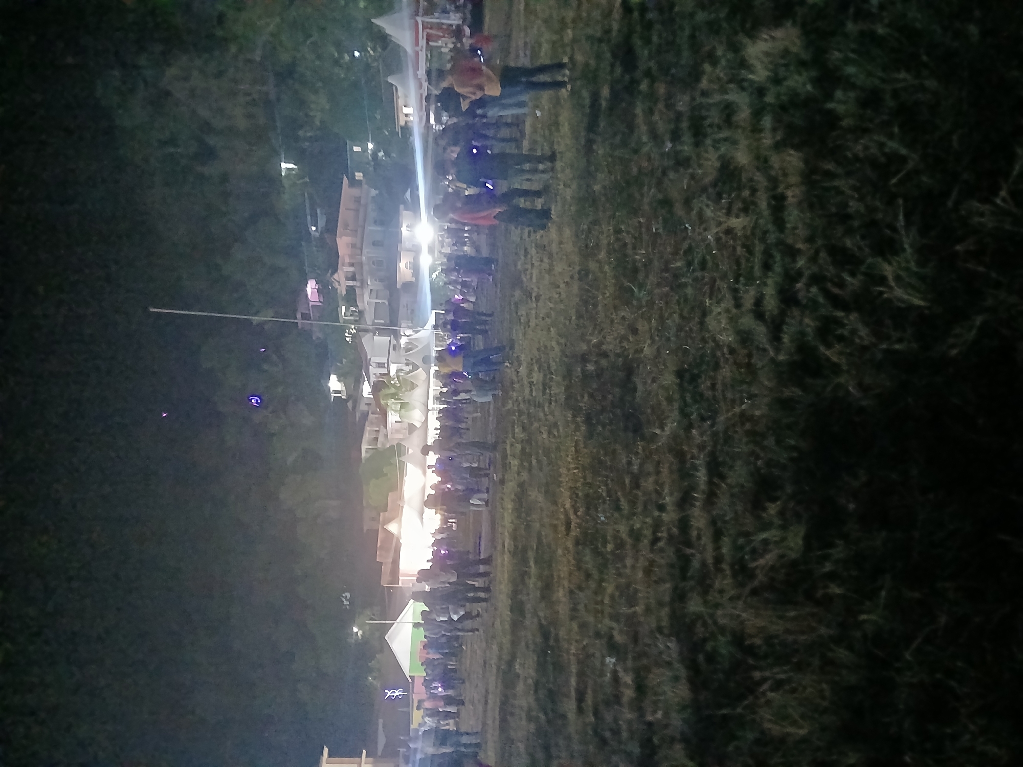 An outdoor evening gathering is depicted, likely a festival or community event, taking place on a grassy field at night. In the background, several buildings with illuminated windows and a hilly, tree-covered landscape are visible. The sky is dark, with a few faint lights and what appear to be two small, glowing orbs.
The foreground is dominated by dry, sparse grass, with shadows cast by unseen light sources. A crowd of people, numbering over fifty, are spread across the field. They appear to be engaged in social interaction, with some standing in groups, others walking, and a few holding small, illuminated objects. Several white tents with red trim are set up in the middle ground, suggesting vendor stalls or activity areas.
The people are dressed in casual clothing, and their faces are largely indistinguishable due to the low light conditions. The overall atmosphere is one of a lively public event occurring after dark in a semi-urban or rural setting. The presence of buildings and infrastructure suggests it is located within the city of Terempa, Indonesia.