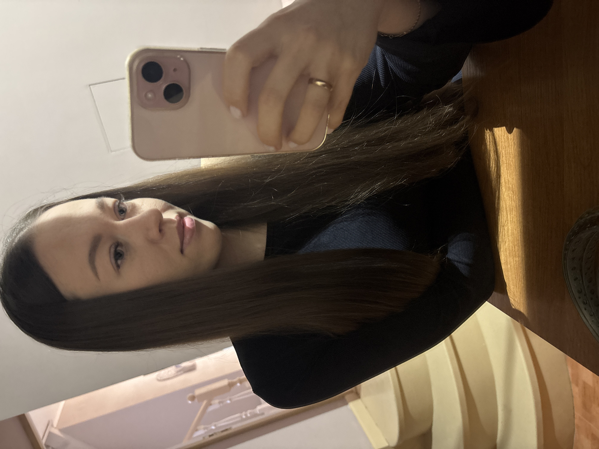 A woman is taking a selfie with a pink smartphone. She is wearing a dark blue top and a gold ring on her left ring finger. She has long, dark brown hair. The photo appears to be taken indoors, possibly in a residence in Ферма, Russia, as indicated by the geotag. The background includes a light-colored wall and a wooden table or surface. Part of what looks like the interior of a room can be seen in the lower left.