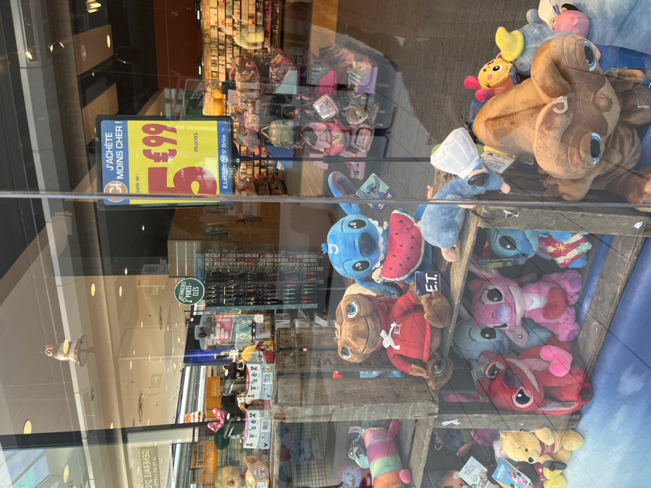 The image shows a storefront display with a variety of plush toys. The storefront is located at 49.283, 4.001 in the city of Saint-Brice-Courcelles, France. The storefront is a store for children's items, such as plush toys, and it is an E.Leclerc store. The display is in the window and is visible from outside. The toys are displayed on wooden shelves, with some plush toys hanging from a rack. The toys are arranged in a random order. Some of the toys are recognizable characters, such as ET and Stitch. The toys are mostly in shades of blue, pink, and brown. The storefront window has a sign advertising plush toys for sale. The sign says "J'achete moins cher!" with a price tag of "5.99."