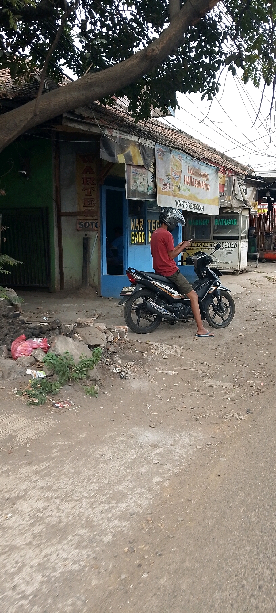 In Cileungsi, Indonesia, a man wearing a red shirt, khaki shorts, a helmet, and flip-flops sits on a black motorcycle parked on a dirt road in front of a small building. The building has a blue door and windows. Signs are visible on the building, one reading "WARTEG BAROKAH" and another partially obscured, with text that appears to be "SOTO". To the left of the building, a green metal gate stands closed. In the foreground, a pile of rocks and debris includes a crumpled red plastic bag and some green foliage. The background features overhead wires and a tree with a thick branch extending over the building.
