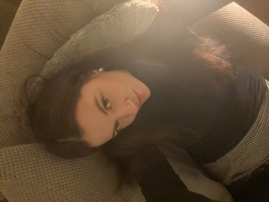 A woman with long, dark hair lies on a light-colored couch with cushions. She wears a black long-sleeved shirt and a patterned skirt or pants. Her head rests on a light green pillow. The lighting is soft, with a bright area in the upper right corner of the frame. The woman looks directly at the camera with a gentle expression.