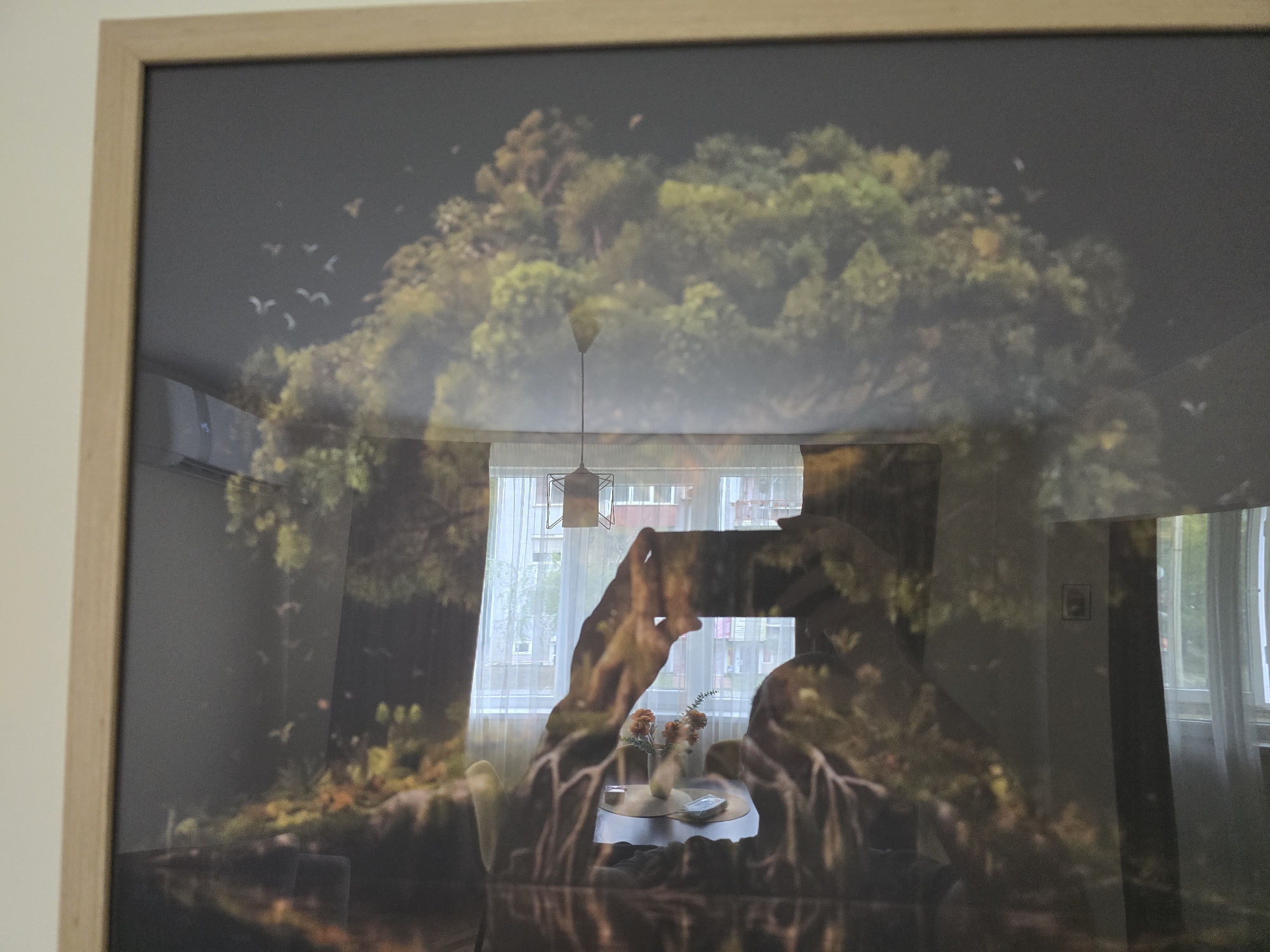 The image displays a framed picture, predominantly featuring a reflection of the room it is in. The frame itself is a light-colored wood.
Within the artwork, a dark, natural scene is depicted. Lush, dense green and brown foliage, likely representing trees or a forest canopy, dominates the background. Above the trees, a dark, muted sky is visible, with several small, white birds in flight on the left side. The foreground of the artwork appears dark, possibly representing water or shadowed ground, with a mirrored reflection effect of the trees and birds.
Superimposed over the artwork, due to the glass reflection, is an interior room scene. A person's hands, holding a smartphone or camera, are clearly visible in the center, indicating they are taking the photograph. Behind the hands, a large window with sheer white curtains allows daylight into the room. Through the window, parts of an external building can be seen. Below the window, a small round table holds a vase with orange/brown flowers and other small objects. An overhead light fixture with a geometric design hangs from the ceiling, also reflected. To the far left, an air conditioning unit is mounted on a light-colored wall. To the right, a small framed picture is visible on a wall.
The scene takes place indoors during the daytime, evidenced by the light filtering through the window. There are no discernible emotions or specific location cues beyond a typical modern indoor setting. No visible text is present.