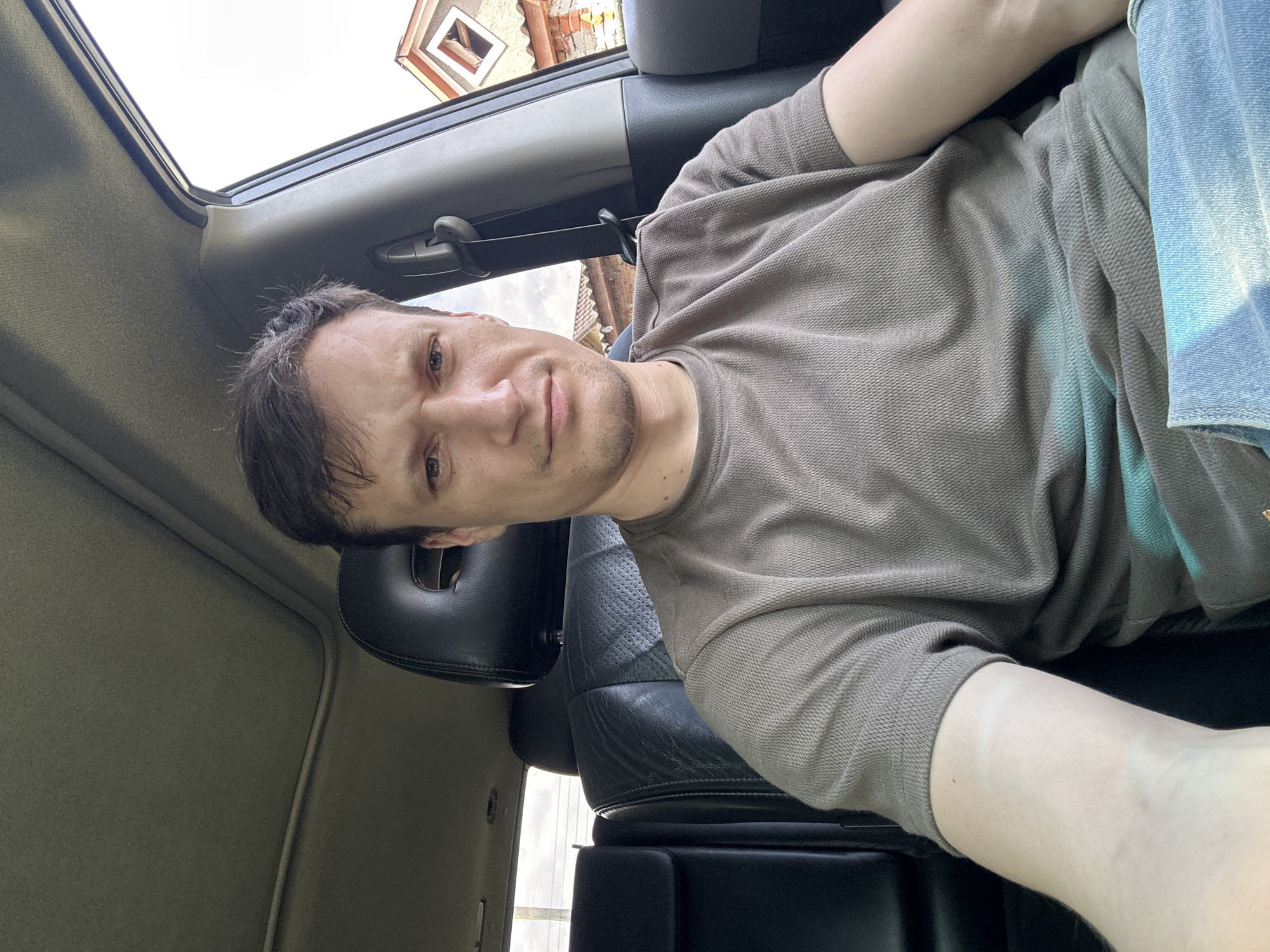 A man with brown hair and a light complexion sits in the driver's seat of a car. He is wearing a grey t-shirt and blue jeans. The car's interior is visible, including black leather seats and a sunroof. Outside the car, a building with a red roof and brick walls can be seen. The scene appears to be in Makiv, Ukraine.