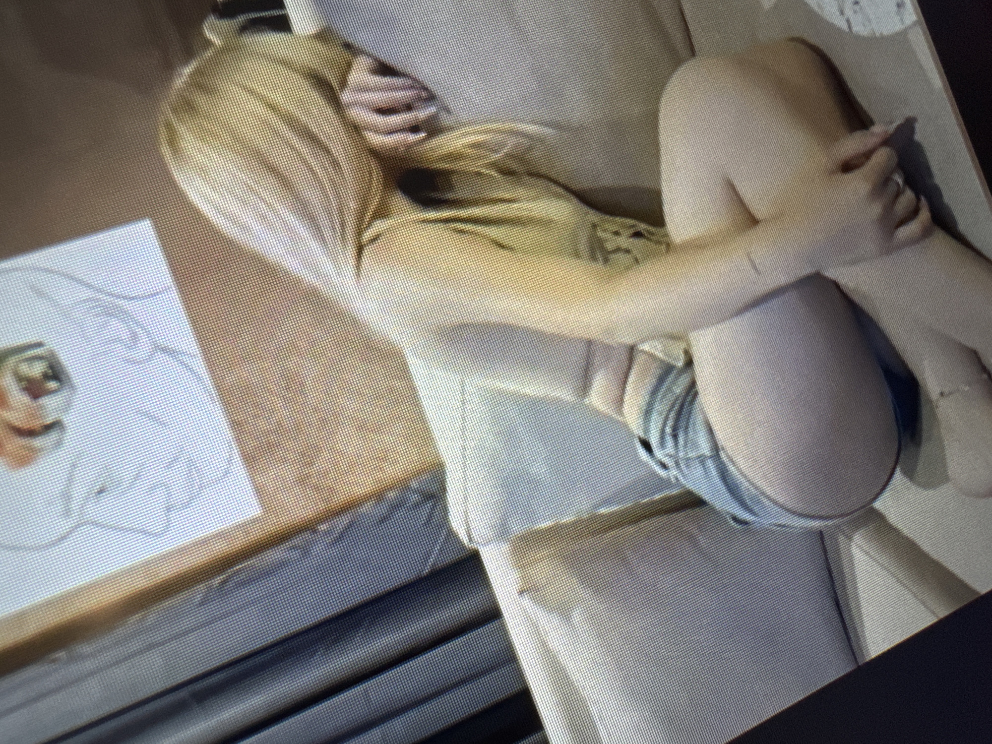 A person with long blonde hair, possibly female, is depicted sitting in a curled-up position on a light-colored sofa or chair. They are wearing a light-colored top and denim shorts, with their arms wrapped around their knees. One hand, adorned with a ring on one finger, is visible resting on their leg.
To the left of the seated person, on a flat surface that appears to be a table or desk, there is a piece of white paper with a line sketch on it, possibly depicting a face or a partial figure. Adjacent to the sketch, a small, partially obscured object with an image on its screen or surface is visible.
The overall image displays a noticeable grid pattern, suggesting it is a photograph of a digital screen rather than a direct photograph of the scene. The person's posture conveys a sense of rest, introspection, or comfort. The setting appears to be indoors. There are no specific cues regarding the time of day, weather, or precise location. No visible text is discernible in the image.