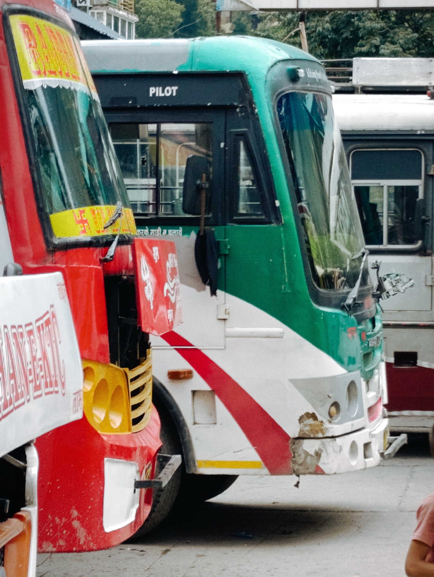 Two buses are parked side-by-side on a street, likely in Manali, India. The bus on the left is predominantly red and white, with a yellow sign featuring text and a swastika symbol. The bus on the right is green and white, with the word "PILOT" visible above its windshield. The front of the green bus shows signs of damage, with chipped paint and exposed material. In the background, the environment suggests an urban or semi-urban setting, with trees and buildings visible. A child's arm, wearing a pink shirt, is partially visible in the lower right corner of the frame, indicating the presence of at least one person observing the scene.