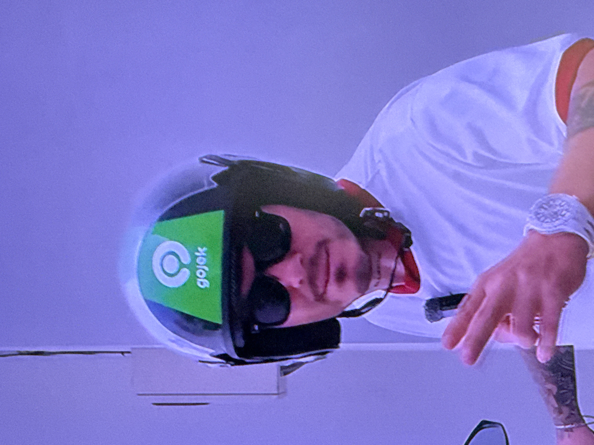 A man wearing a white tank top, sunglasses, and a black helmet with a green Gojek logo is on what appears to be a motorcycle or scooter. The background is a plain, light purplish color with a horizontal line near the bottom. There is no indication of Bietigheim-Bissingen, Germany, in the image. The man is holding the handlebars of the vehicle with a watch on his left wrist. He has visible tattoos on his arm.