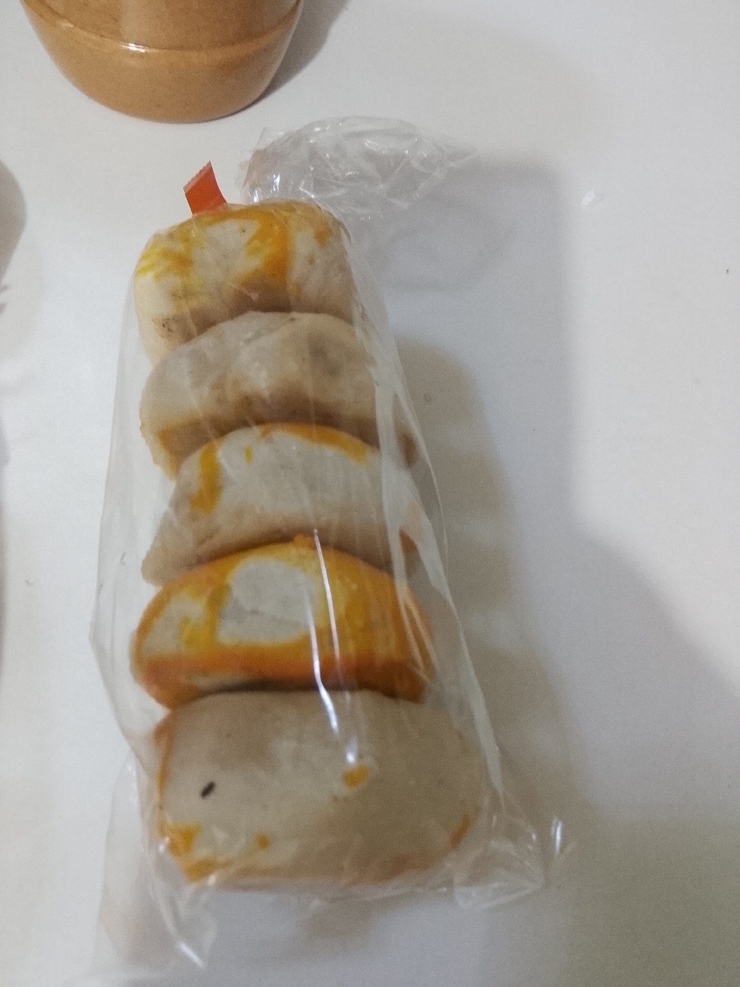 The image displays five individually formed, stacked pastries or snacks enclosed within a transparent plastic bag, observed within an interior setting documented as Davao City, Philippines. The pastries exhibit a pale beige or off-white base color with distinct, irregular bright yellow-orange streaks or glaze visible on their exposed surfaces, particularly along their edges. They possess a rounded, somewhat crescent or semi-oval shape. The plastic bag is secured at the top by a small, folded orange tab. A minor, dark, punctiform object is present on the surface of the lowest visible pastry. The bag rests on a flat, white, untextured surface. A segment of a light brown, curved object, resembling a bowl or container, is partially visible in the upper left background. The scene is brightly and uniformly illuminated, casting minimal shadows.