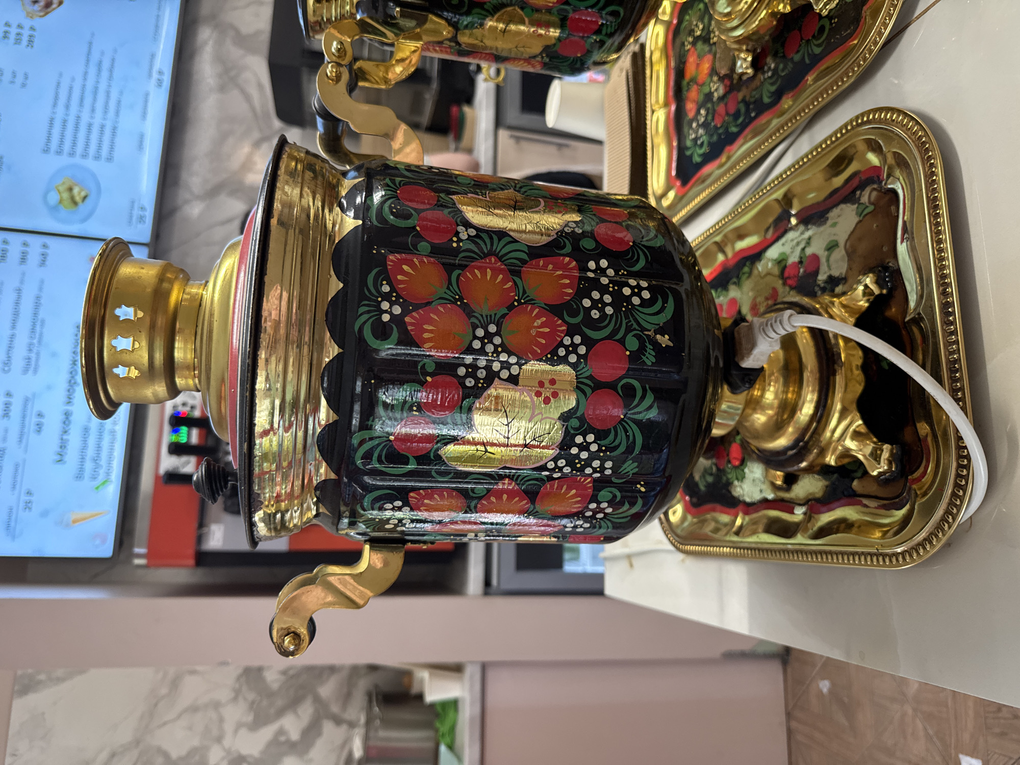 This media file depicts an ornate, traditional Russian samovar set in an indoor cafe or eatery, likely in Saint Petersburg, Russia. The samovar, a central feature, is richly decorated with a black base and vibrant folk art painting. It showcases red and orange floral or berry motifs, green leaves, and white dot patterns, highlighted by striking gold gilded elements that resemble stylized foliage. Its metallic components, including the top, spigot, handles, and feet, are made of gleaming gold-colored metal. A white electrical cord visible at the back indicates it is an electric model.
The samovar stands on a decorative, gold-colored rectangular tray, which itself features subtle painted patterns. Another similar, partially visible tray is situated behind it. In the background to the left, a digital menu screen displays items and prices in Russian. Notable text includes "Сбитень подовый 100", "Мягкое мороженое" (Soft ice cream) with "Ванильное" (Vanilla) and "Клубничное" (Strawberry) flavors, and several "Блинчик" (pancakes or crepes) options such as "с творогом" (with cottage cheese), "с вишней" (with cherry), and "с яблоком" (with apple). Various prices, including "25 P", "40 P", "159 P", and "289 P", are listed. The presence of the samovar and the menu indicates a setting for serving beverages, possibly traditional tea, and casual meals. No people are visible, and no specific event is occurring beyond the presentation of these items. The image captures an indoor scene, so time of day or external weather conditions are not apparent.