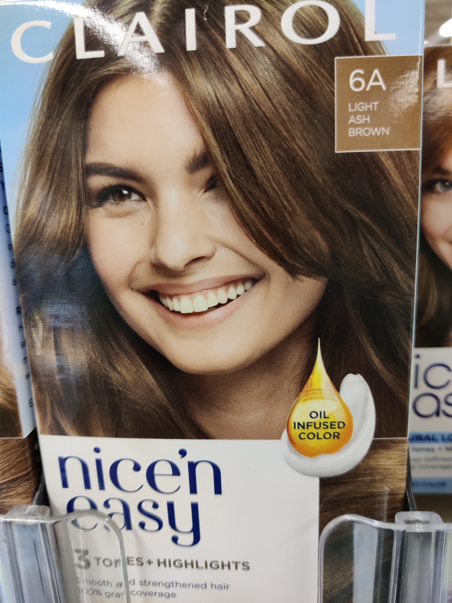 A Clairol Nice'n Easy hair color box, shade 6A Light Ash Brown, is displayed in a retail setting in Mechanicsburg, United States of America. The box features a close-up image of a smiling woman with brown hair, her eyes are looking directly at the viewer. Text on the box includes "CLAIROL," "nice'n easy," "6A LIGHT ASH BROWN," and "OIL INFUSED COLOR." The product is positioned in front of other similar products, partially obscuring them. The lighting appears to be from overhead retail lights.