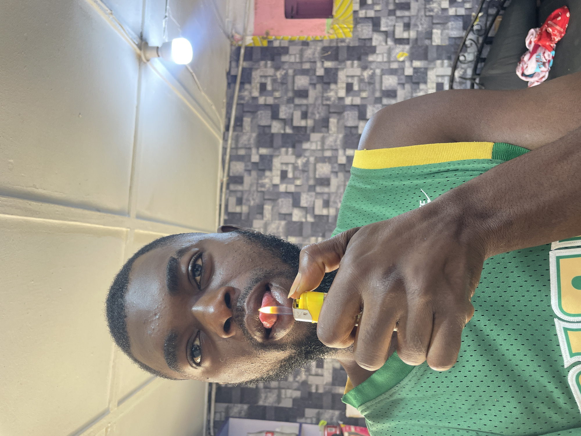 The image captures a young, dark-skinned man in an indoor setting in Jos, Nigeria. He has a full beard and short dark hair, and his eyes are wide and focused, looking directly at the camera. He is wearing a green and yellow mesh sports jersey, with a distinct perforated texture visible.
The central activity shows the man holding a yellow plastic lighter in his right hand. The lighter is ignited, and a small, bright orange flame is visible, positioned very close to or touching his tongue, indicating he is performing a fire stunt or trick.
The background reveals details of the room. To the left (above the man's head in the image's orientation) is a light-colored wall with clear paneling or seams, featuring a bright, unshaded white LED light bulb mounted on it. Behind the man, further to the right, is a wall adorned with a grey and black geometric patterned wallpaper. In the far right background, a dark surface, possibly part of a chair, holds a crumpled red and white piece of cloth or garment. The lighting in the scene is artificial, primarily from the visible bulb. No legible text is visible in the image.