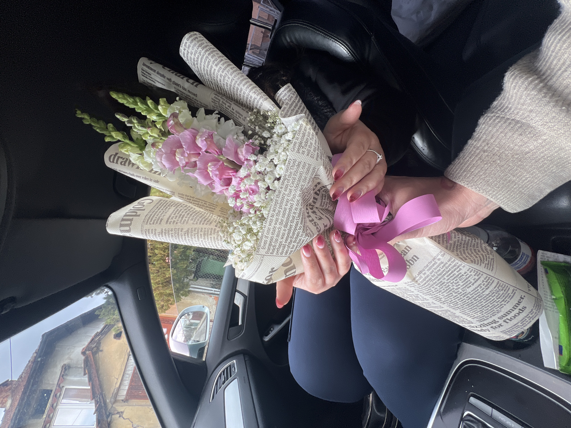 An overhead shot inside a dark-interior vehicle captures two individuals' hands holding a floral bouquet. The bouquet features pink and white snapdragons, white baby's breath, and green foliage, wrapped in paper resembling newspaper print and secured with a bright pink ribbon. The upper pair of hands, with manicured nails displaying a red-to-pink gradient, wears a silver ring with a clear stone on the left ring finger and belongs to a person in dark blue attire. These hands hold the bouquet's midsection. The lower hands, partially visible and wearing a light-colored, ribbed knit sweater, support the bouquet from below, indicating an exchange or shared holding.
The car's interior shows a dashboard segment, an air vent, a door handle, and part of the center console with an "ESP OFF" button, a clear plastic bottle, and a green wrapper. Through the front passenger window, an external side mirror reflects a street scene. Outside, an older two-story building with a reddish-brown tiled roof, light-colored stucco walls, and visible cracks on its facade is present. Wooden shutters adorn some windows. Green trees and bushes are visible beneath an overcast sky. This architectural and urban setting is consistent with a residential area in Sofia, Bulgaria.