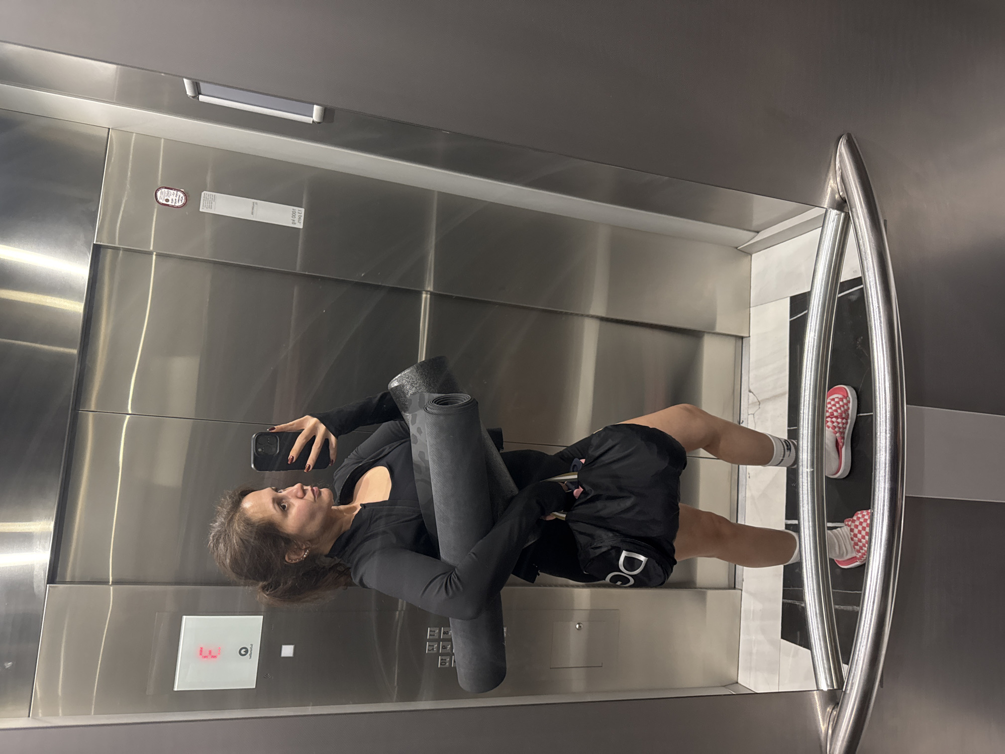 A mirror selfie captures a woman inside a modern elevator with brushed stainless steel walls. The woman, viewed from the chest up and facing left, has medium-length dark hair and wears a black long-sleeved athletic top, black athletic shorts with partial white lettering (appearing as "DO" or "DG"), white socks, and distinctive red and white checkered athletic shoes. She holds a smartphone with red-painted nails in her left hand, appearing to take the photo.
She is carrying two rolled dark-colored mats, possibly yoga mats, one of which exhibits a subtle leopard print pattern. The elevator's control panel, visible to her left, displays a red illuminated "F" and a down arrow, possibly indicating a "Fitness" floor. Another small red indicator light and an unreadable white label are situated higher on the wall. A polished metal handrail is visible on the right side of the elevator, reflecting the black and white tiled floor. The overall setting suggests an urban or hotel environment, consistent with the provided location context of Dubai, United Arab Emirates. The activity is a casual selfie, likely before or after a workout session.