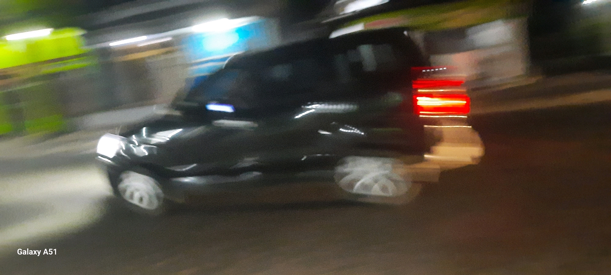 The image, captured with a Samsung Galaxy A51, depicts a moving black SUV at night, likely in Selopuro, Indonesia, due to the timestamp. The vehicle's motion blur is pronounced, with streaks of light indicating movement along a road. The SUV's headlights are illuminated, casting a bright beam forward, while its taillights glow red, suggesting it is either braking or its lights are on. The surrounding environment appears to be an urban or semi-urban area, with blurred streaks of neon green and blue lights suggesting buildings or signs in the background. The road surface is dark and indistinct due to the motion. No people are clearly visible within or around the vehicle. The overall impression is a dynamic night scene of a vehicle in transit.