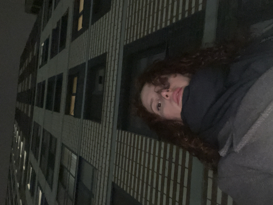 A low-angle, nighttime photograph depicts a female subject in the foreground, positioned in Saint Petersburg, Russia. The female has long, dark brown, curly hair and is wearing a dark overcoat and a dark scarf that covers her chin and lower face. Her visible facial features include her nose, eyes, and pink lips, and she gazes directly at the camera. In the background, a tall, multi-story building with a light-toned brick facade and dark, rectangular window frames rises steeply into the dark sky. Several windows are illuminated with warm, yellow light, while others appear dark. The precise location within Saint Petersburg is not identifiable from the visible details.