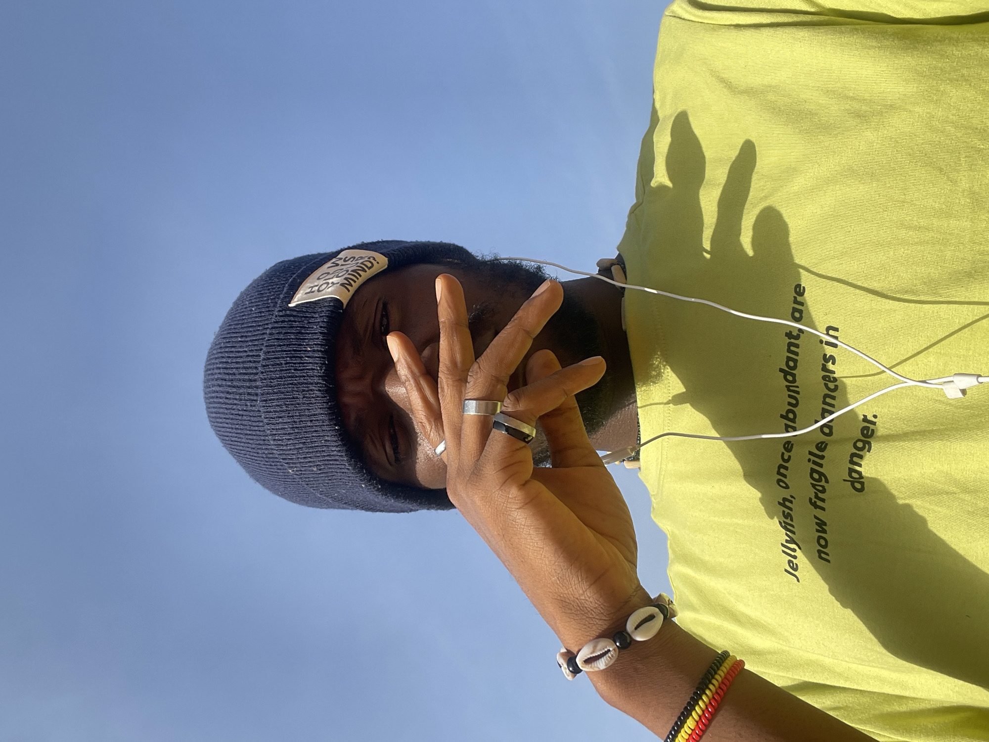 A low-angle shot of a dark-skinned person wearing a navy blue beanie with a white tag, a light green t-shirt with text that reads "Jellyfish, once abundant, are now fragile dancers in danger," and white wired earphones. The person has rings on their fingers and bracelets on their wrist. The background is a clear blue sky. There is no information available in the image to determine if this image was taken in Jos, Nigeria.