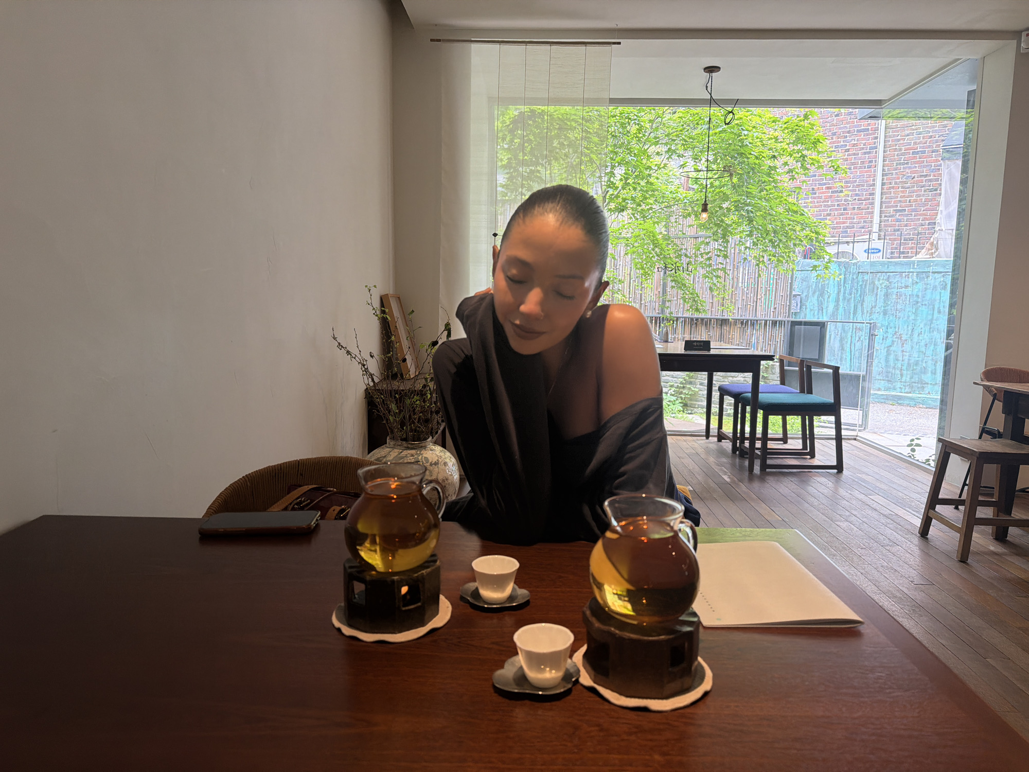 A woman with dark hair pulled back is seated at a wooden table in what appears to be a modern tea house or cafe. She wears a dark, off-the-shoulder garment and is looking down with a serene expression, her eyes closed or cast downwards.
On the dark wooden table in front of her are two glass teapots filled with amber-colored tea, each resting on a dark, hexagonal stand, possibly containing a warmer. Beside each teapot are two small, white ceramic cups on dark saucers, also resting on white coasters. A smartphone lies face down on the table to her left, near a woven-back chair. Further to the left, a decorative vase with dried branches is visible.
The setting is brightly lit by natural light streaming through a large, floor-to-ceiling window that dominates the right side of the frame. Outside the window, vibrant green foliage of trees is visible, suggesting a warm, sunny day. Beyond the trees, there's a light-colored brick building and a textured, turquoise wall or fence. A black table with blue-cushioned chairs is seen through the window, inside the cafe, suggesting more seating. A single pendant light with an exposed bulb hangs near the window. The floor inside the cafe is made of dark wooden planks.
On the table near the right teapot, a white booklet or menu is placed. Visible Korean text includes "예약석" (reservation seat) on a small sign outside the window and some characters on the menu/booklet. The overall ambiance is calm and contemplative, reflecting a traditional tea-drinking experience in a contemporary setting in Seoul, South Korea.