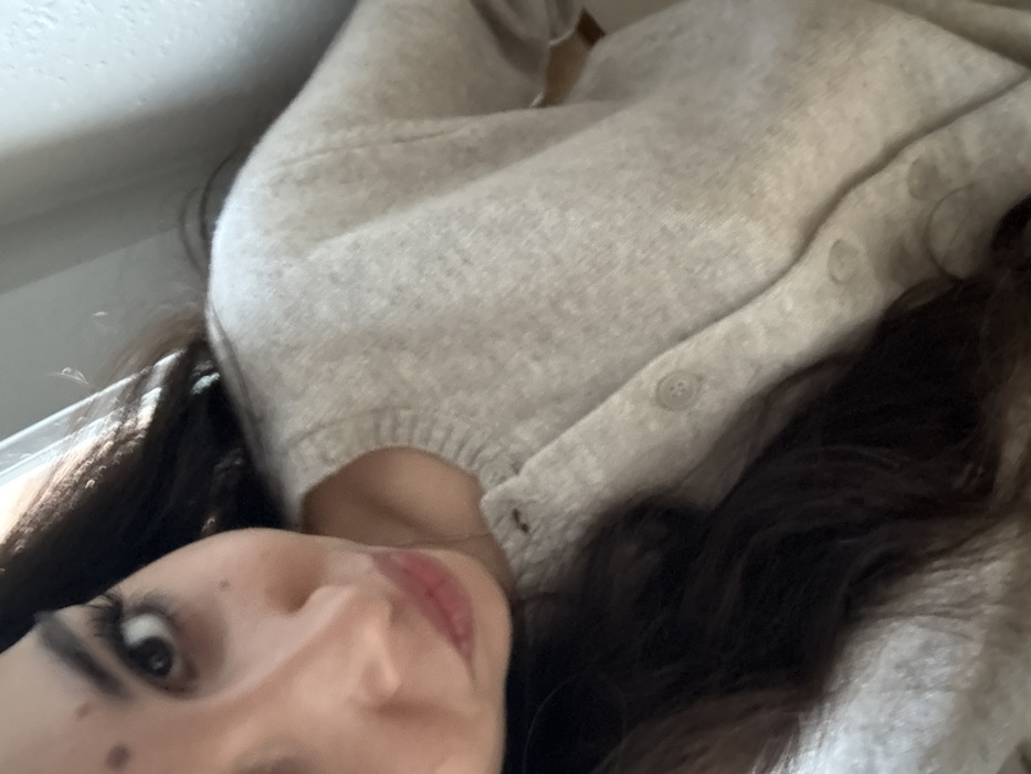 A woman lies on her side, her face close to the camera. She has dark hair and wears a light-colored, button-down sweater. Her expression is soft, and her lips are slightly parted. The background appears to be a room with natural light coming in from a window. The photo's angle is tilted, giving a casual, intimate feel.