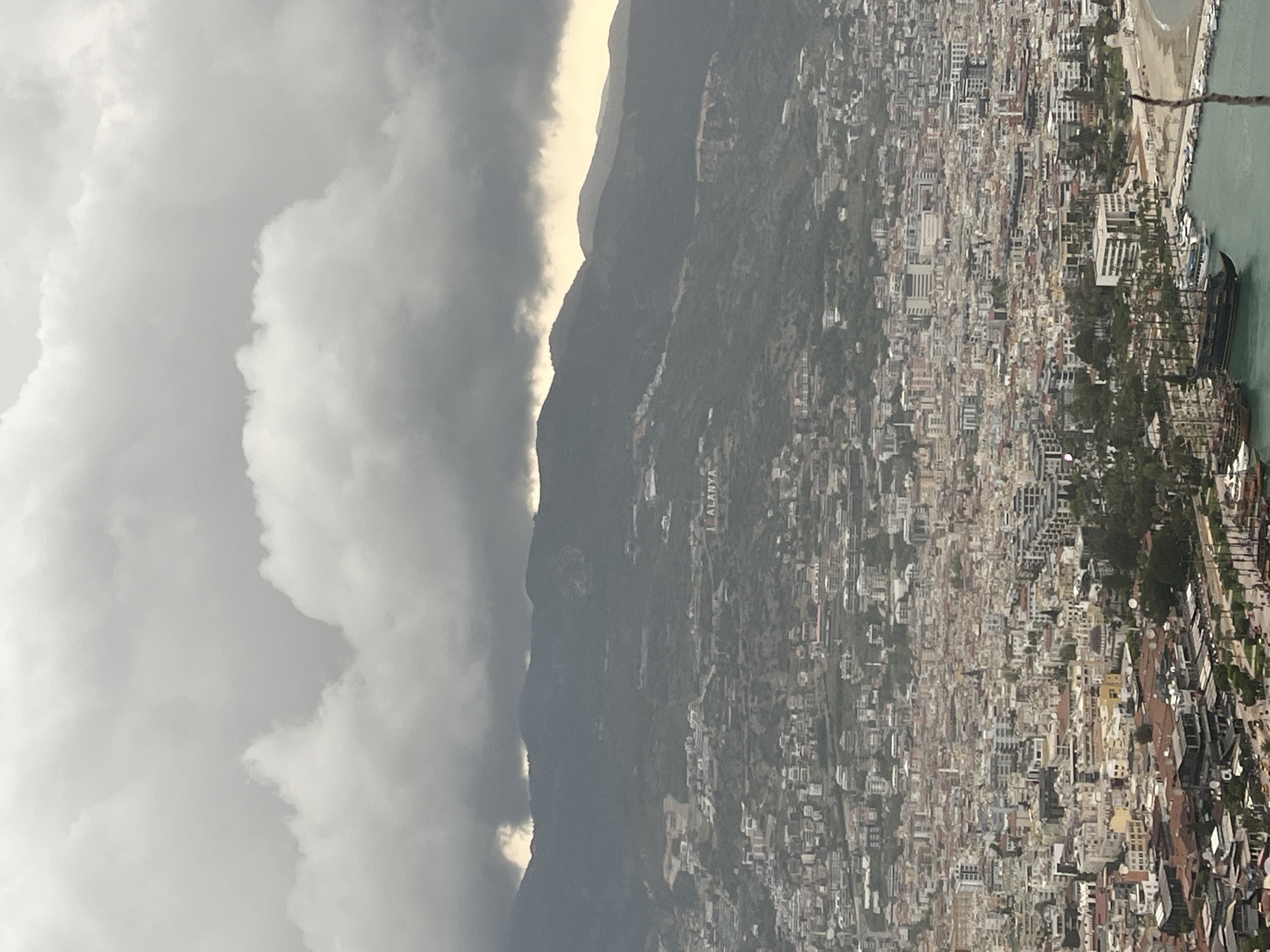 The image displays an expansive, high-angle view of the coastal city of Alanya, Turkey, situated between a mountainous backdrop and the Mediterranean Sea, under a heavily clouded sky.
At the top of the frame, dense, dark grey clouds dominate the sky, partially obscuring the upper slopes of the mountain range. A brighter strip of sky is visible just above the mountain peaks, suggesting either filtered sunlight attempting to penetrate the cloud cover or a break in the clouds further in the distance.
The middle section features a prominent range of dark, rugged mountains, covered with vegetation, their slopes descending towards the urban area. On one of these hillsides, large white letters distinctly spell out "ALANYA," clearly identifying the location.
The lower half of the image is filled by the densely populated cityscape of Alanya. Numerous buildings, predominantly light-colored with some red-tiled roofs, are closely packed together, stretching from the base of the mountains to the shoreline. Green patches of trees and vegetation are interspersed throughout the urban landscape.
Towards the bottom right, the scene transitions to a narrow sandy beach bordering the calm, light blue-green waters of the Mediterranean Sea. Several boats are visible near the coast, including a large, dark, multi-masted ship, possibly a replica pirate ship used for tourism, docked in the bay. The weather appears overcast and moody due to the extensive cloud cover, suggesting either a cloudy day or a time near sunrise or sunset.