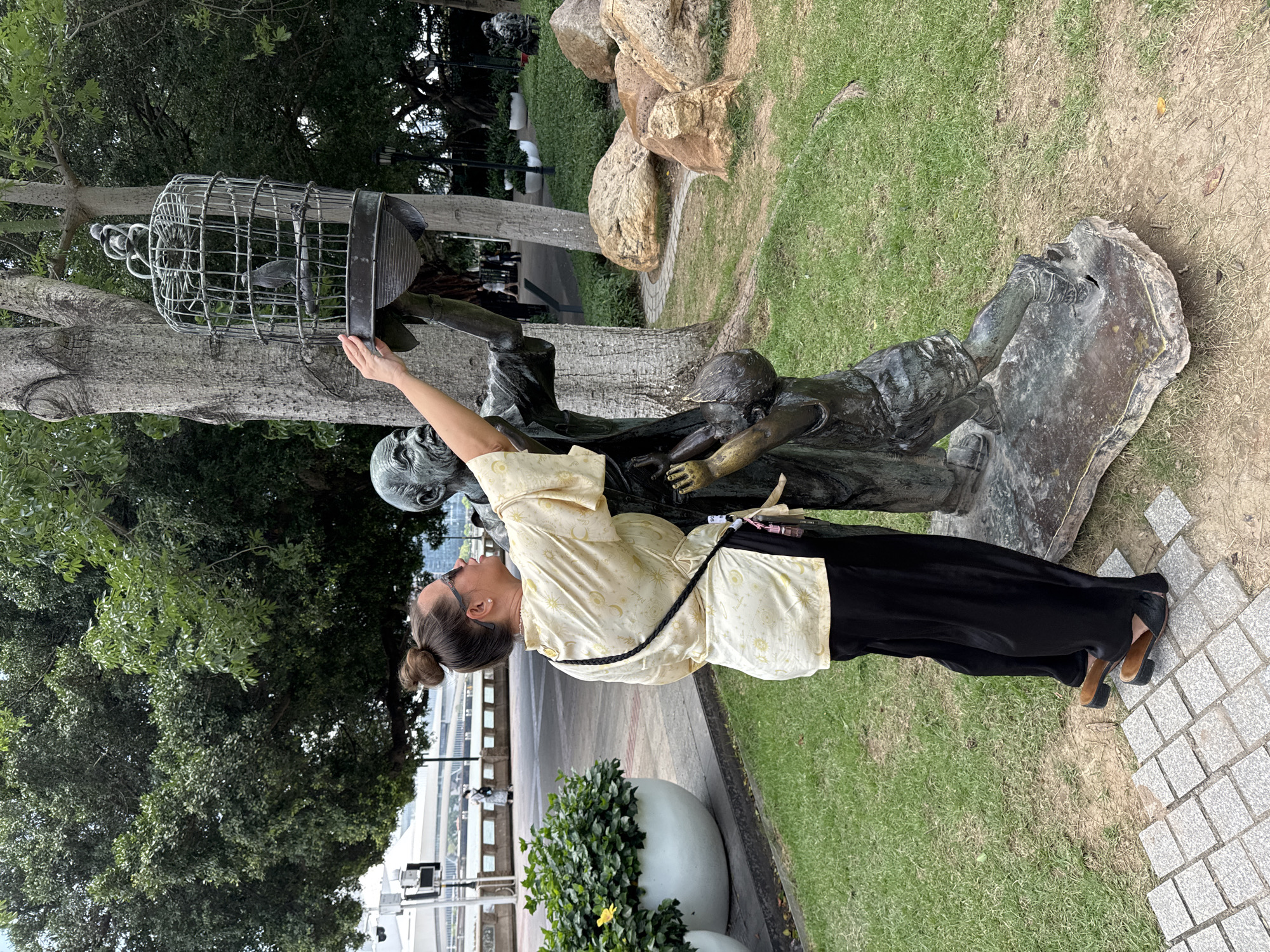 A woman stands in front of a bronze statue in a park, likely in the Liwan District, China. She is wearing a light-yellow, patterned top and black pants with brown shoes. Her hair is pulled back in a bun. Her right arm is extended, appearing to touch a birdcage held aloft by the bronze figures. The statue depicts two figures hoisting the cage. The environment includes green grass, trees, paved walkways, and large rocks. In the background, there's evidence of urban development.