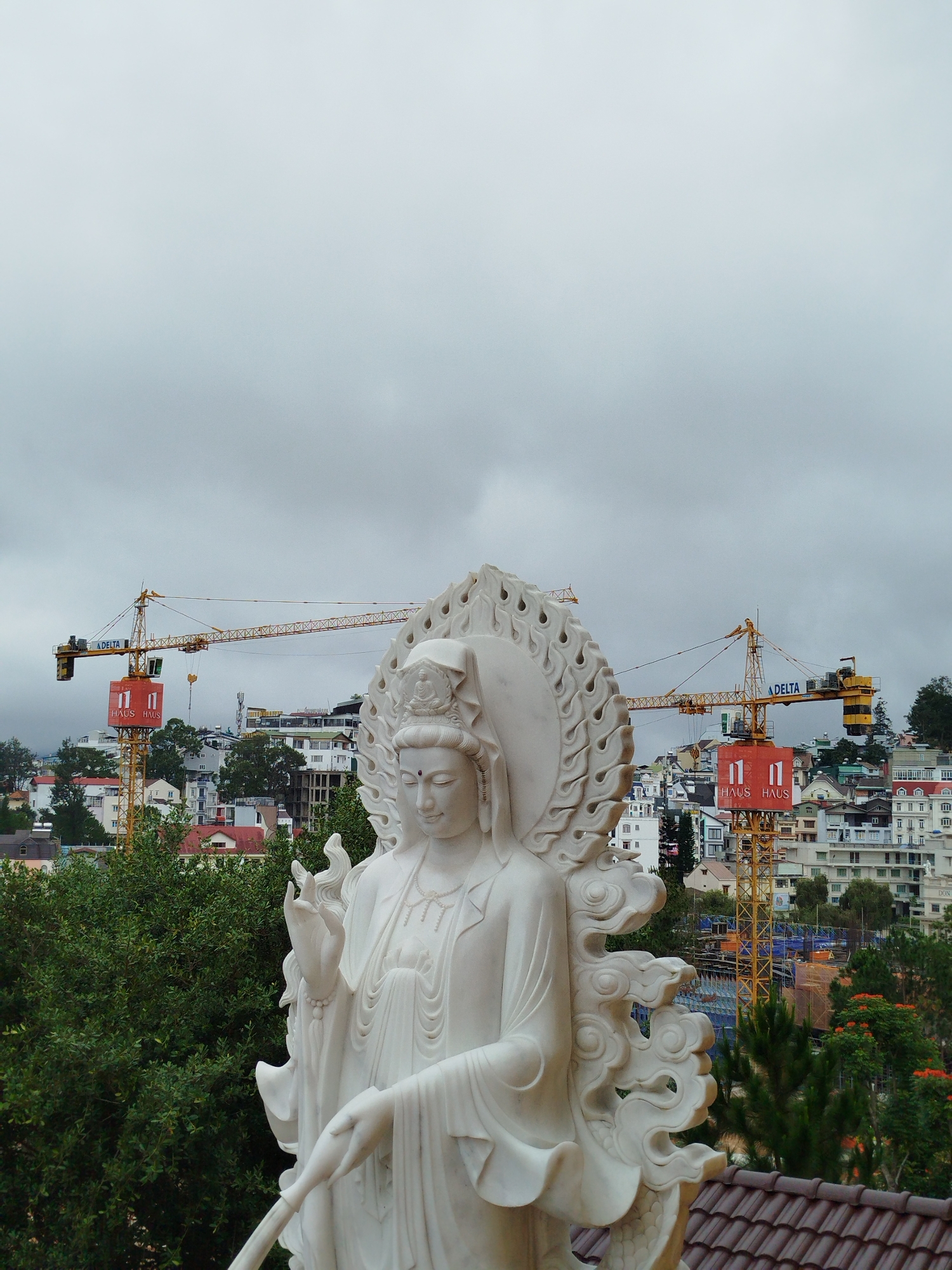 A white marble statue of a Buddhist deity, possibly Guan Yin, stands in the foreground. The deity is depicted with multiple arms and is adorned with intricate robes and jewelry. The statue is positioned on a rooftop in the city of Da Lat, Vietnam, with a view of the surrounding cityscape. In the background, several yellow construction cranes are visible, indicating ongoing development in the area. Buildings of various sizes and architectural styles are spread across the landscape, interspersed with green trees. The sky is overcast, contributing to a subdued atmosphere. The overall scene captures a juxtaposition of traditional religious art with modern urban construction in Da Lat.