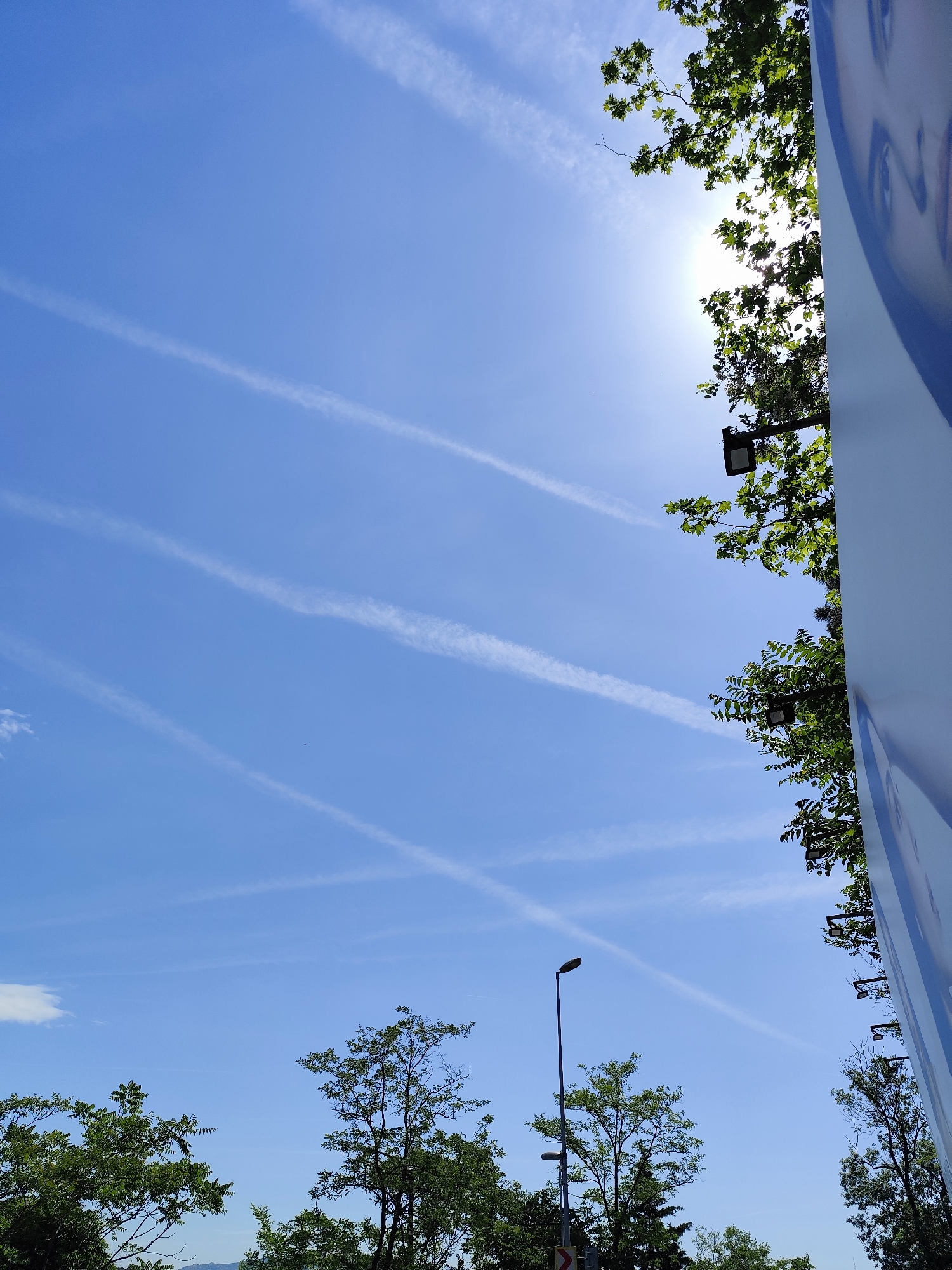 The image displays a bright blue sky on a sunny day, featuring several prominent white contrails crisscrossing each other. The sun is visible in the upper right corner, partially obscured by green tree leaves, creating a strong lens flare.
Green trees with lush foliage frame the bottom and right side of the image. On the far right, a tall, vertical light blue billboard or sign shows a partial, abstract depiction of a person's face, specifically an eye and part of the forehead, rendered in lighter tones. Several black streetlights are visible; one stands centrally in the lower-middle-left, while others are integrated among the trees on the right.
A small, dark speck, possibly a bird, is seen in the middle-left area of the sky. In the lower-left, a small white cloud is present, and faint distant mountains or hills are visible below the trees. A small white road sign with a red left-pointing chevron is partially visible near the base of the central streetlight.
The scene depicts a bright, clear day, likely midday or early afternoon, in an outdoor setting in Sarıyer, Turkey, characterized by both natural greenery and urban fixtures. No discernible text is visible in the image.