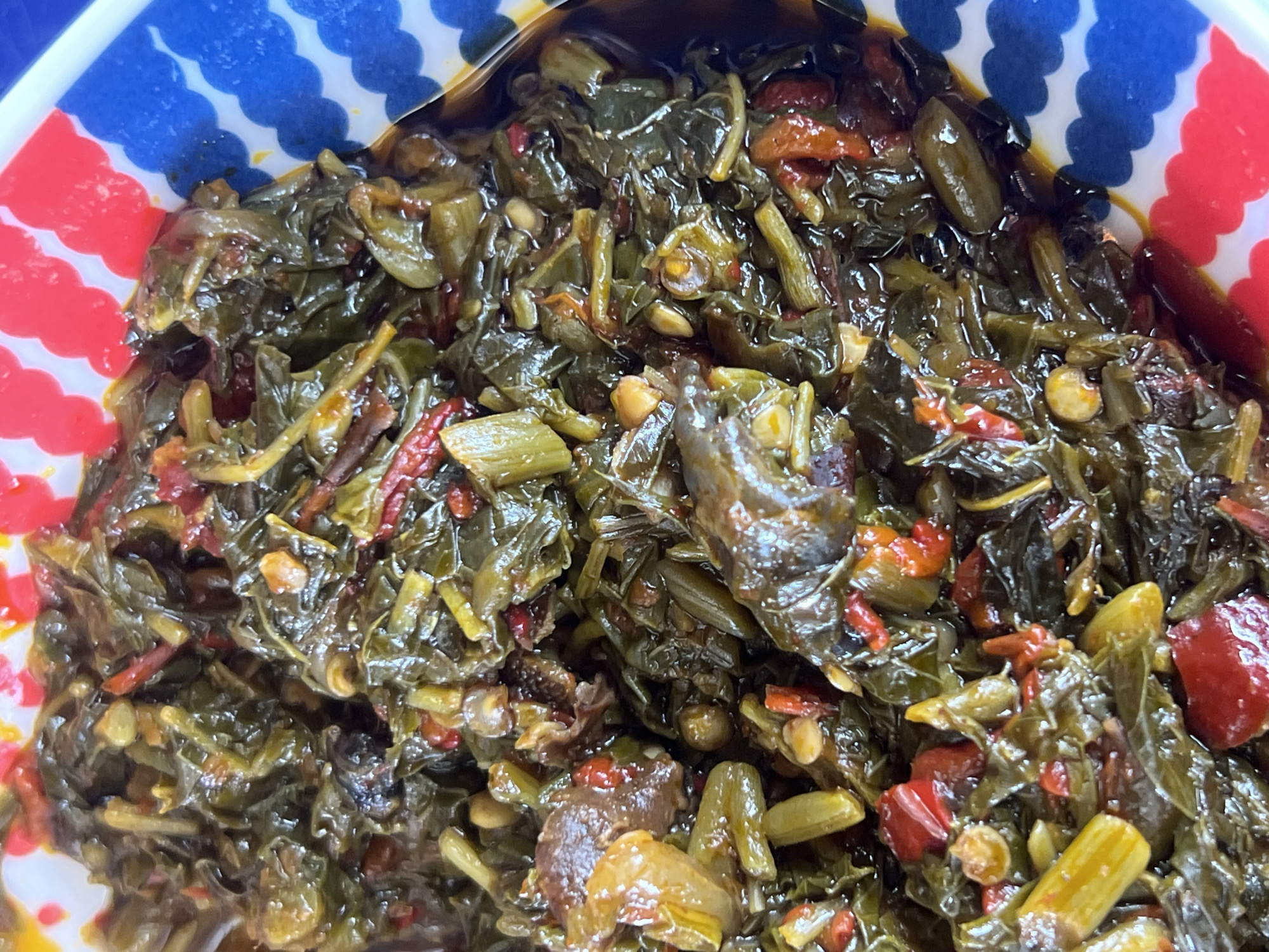 A close-up, top-down view of a cooked dish is presented, identified as being from the city of Jalingo, Nigeria. The primary subject is a dark green, leafy vegetable stew or preparation, appearing moist and glistening. Interspersed within the greens are irregular reddish-orange components, possibly peppers or tomatoes, and smaller, elongated, pale yellowish-green elements, which could be vegetable stems or specific additions like okra or beans. Additionally, some darker, irregularly shaped pieces are visible, potentially indicating protein such as meat or dried fish. The food is contained within a white bowl, the rim of which is partially visible along the top and right side. This rim features a distinct decorative pattern of alternating red and blue scalloped stripes. No individuals, specific actions, or further environmental details beyond the dish and its immediate container are discernible in the frame.