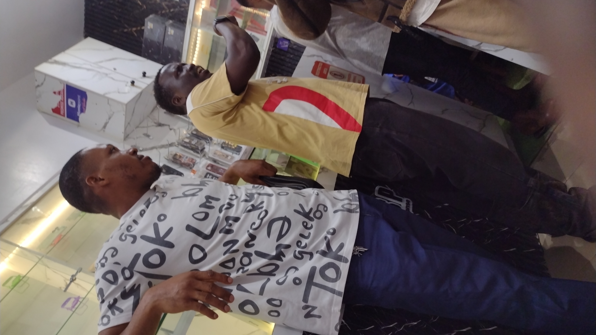 The image depicts two men standing in what appears to be a retail environment. The man closest to the camera is wearing a white t-shirt with black lettering and blue pants. He is looking upwards. Behind him, the second man is wearing a yellow t-shirt with a red and white design, along with dark pants. He is looking upwards as well. The location includes shelving with various small items, possibly phone accessories. A marble-patterned counter is visible, and there are other individuals partially visible in the background. The city and specific location are unknown.