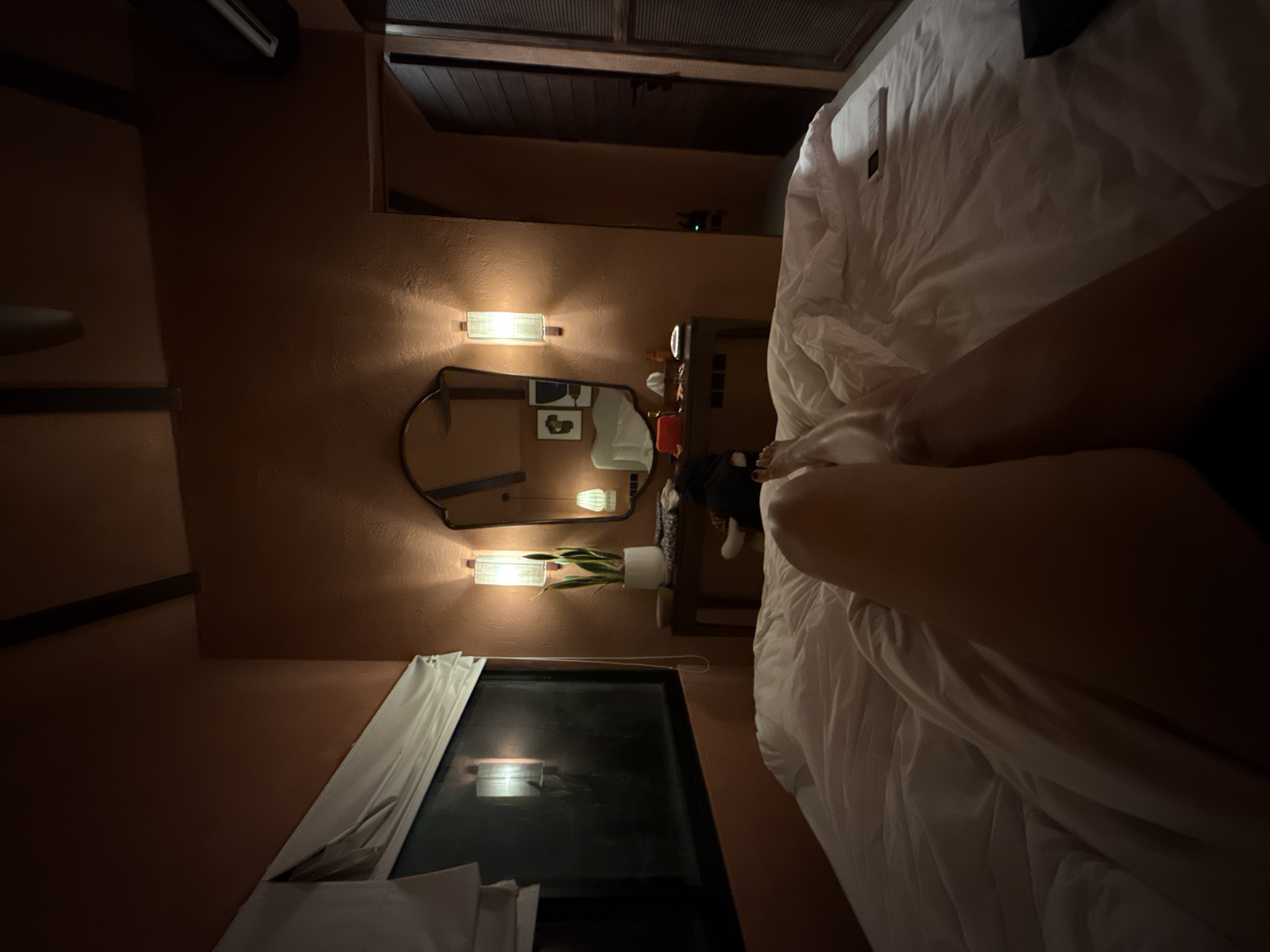A nighttime interior view in Kuta Selatan, Indonesia, from the vantage point of a bed covered with white bedding. Visible are the legs of a person lying in the bed. On the wall is a television, closed curtains, a mirror with two sconces, and a small desk with a potted plant.