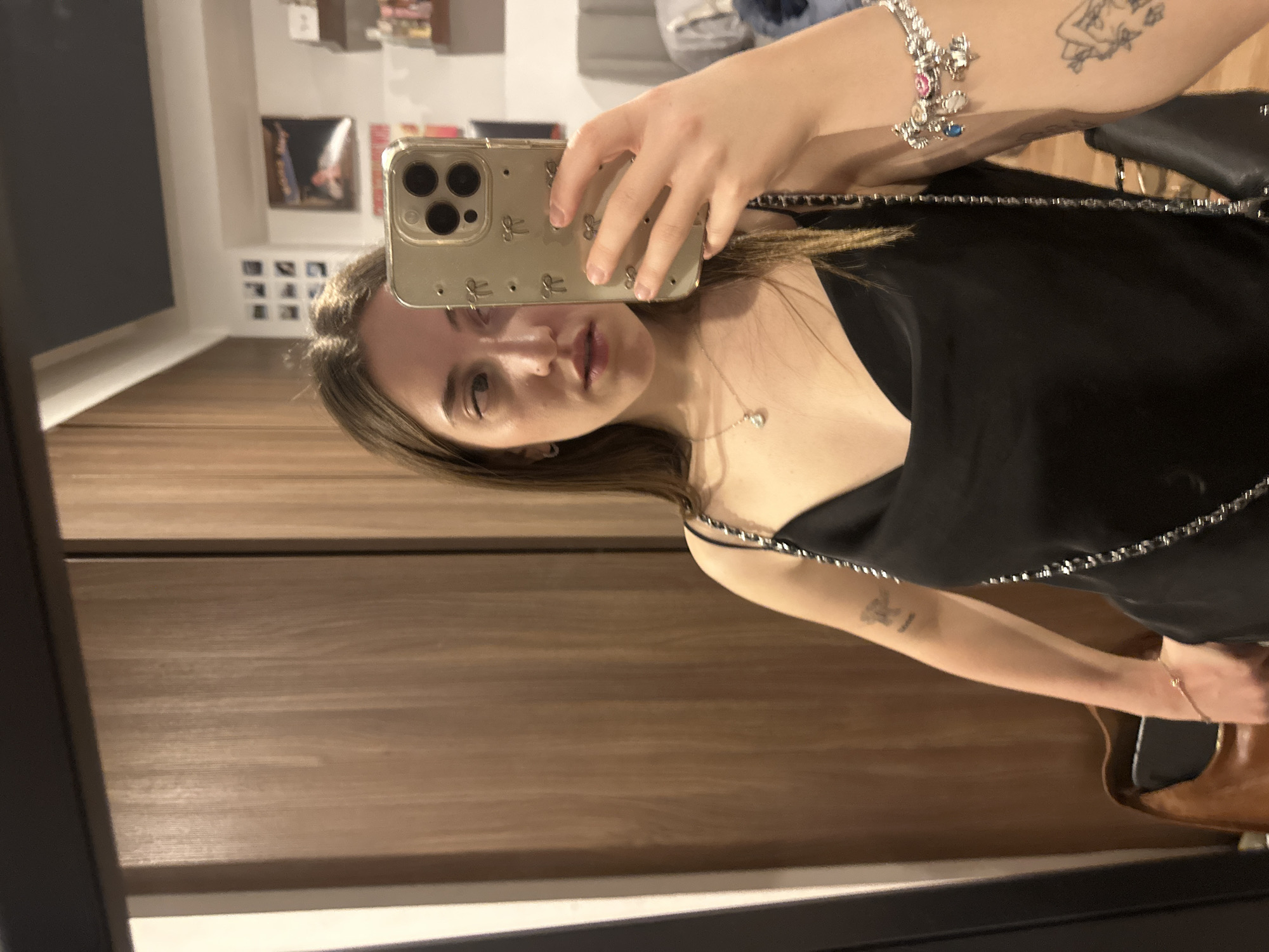 A young woman is taking a mirror selfie inside a building. The phone is gold-colored with a case decorated with bows. She is wearing a black sleeveless dress with chain straps and a delicate necklace. Her left arm is visible, adorned with a bracelet and a tattoo. The background includes a wooden cabinet and a glimpse of shelves and other items in the reflection. The location is likely in Huixquilucan de Degollado, Mexico, but the specific building or street cannot be determined from this single image.